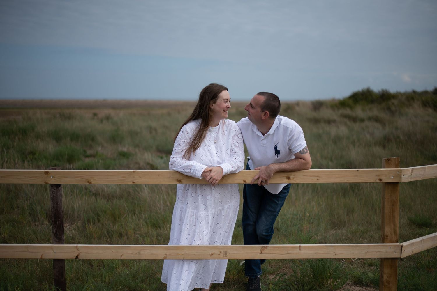 Couple leaning on wooden fence smiling at each other in grassy coastal field.