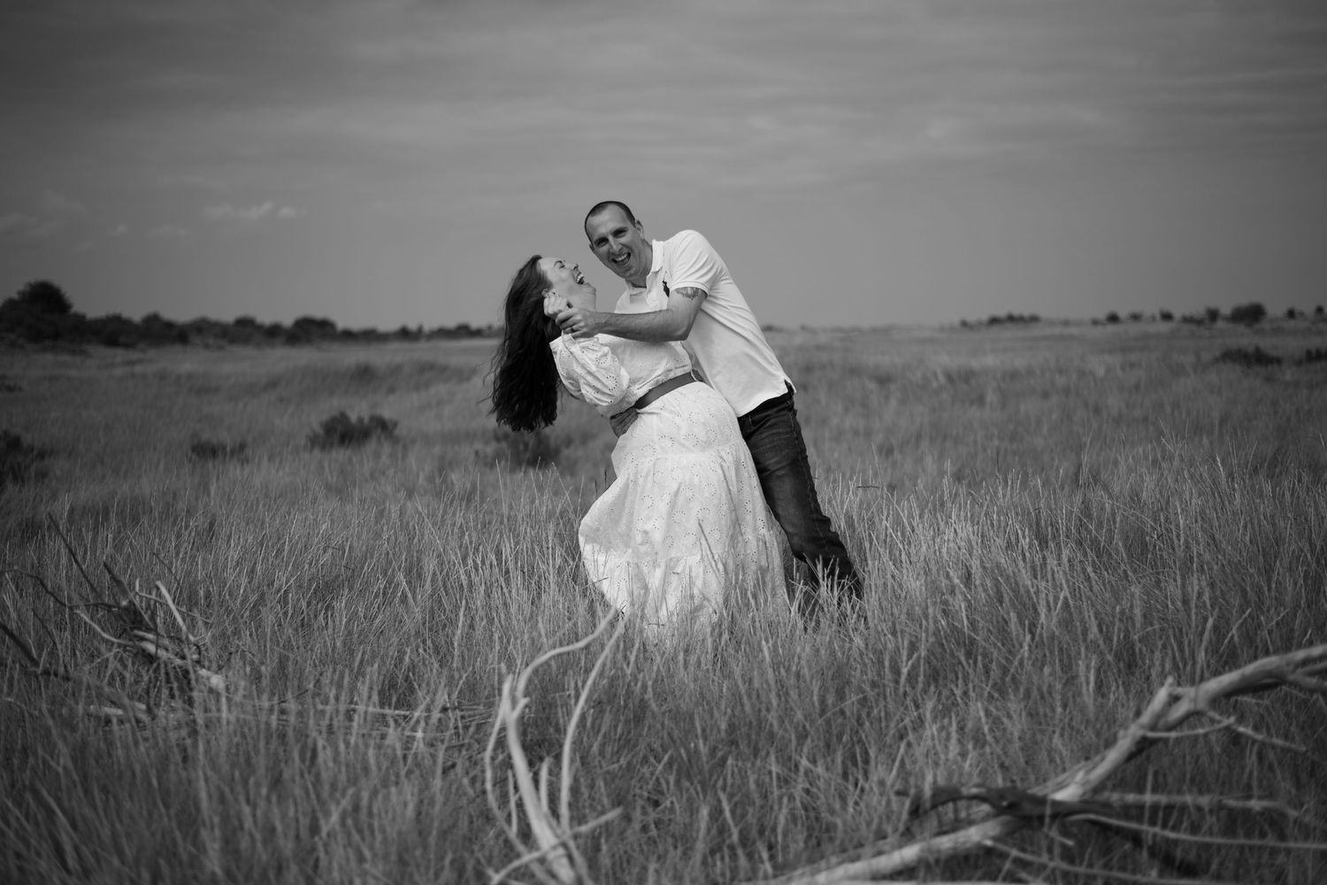 Black and white photo of couple kissing passionately in open grassland field, woman in flowing white dress.