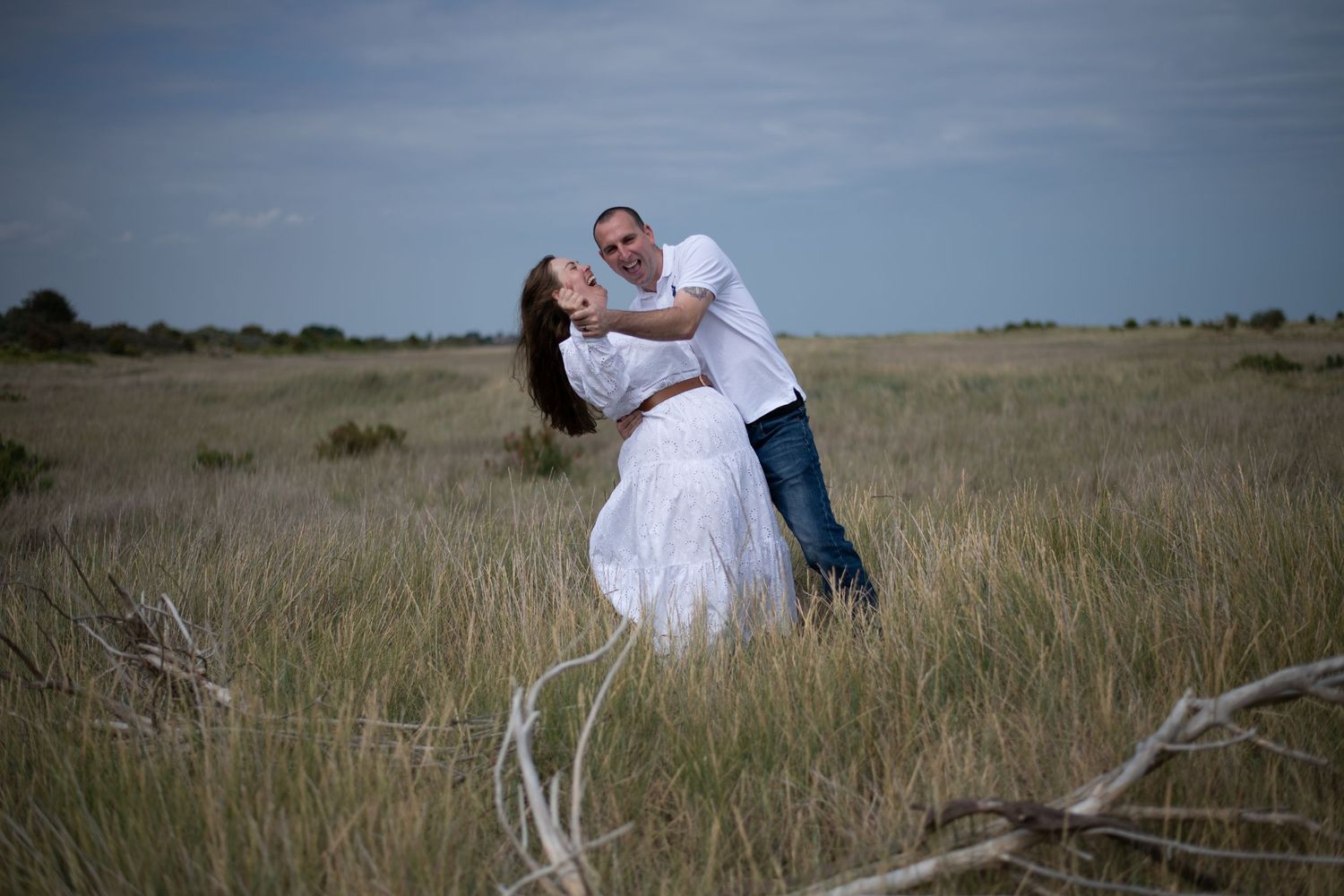 Couple sharing a romantic dip kiss in a wide open grass field, woman wearing a flowing white dress outdoors.