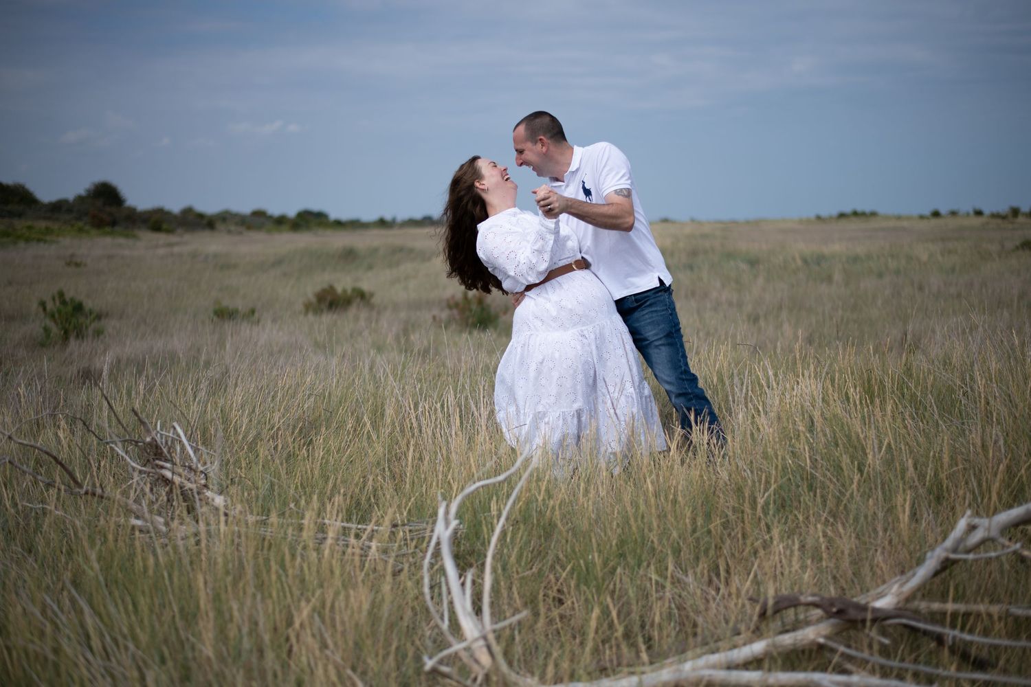 Romantic couple sharing a kiss in a golden grass field, woman in white dress, man in jeans and white shirt.