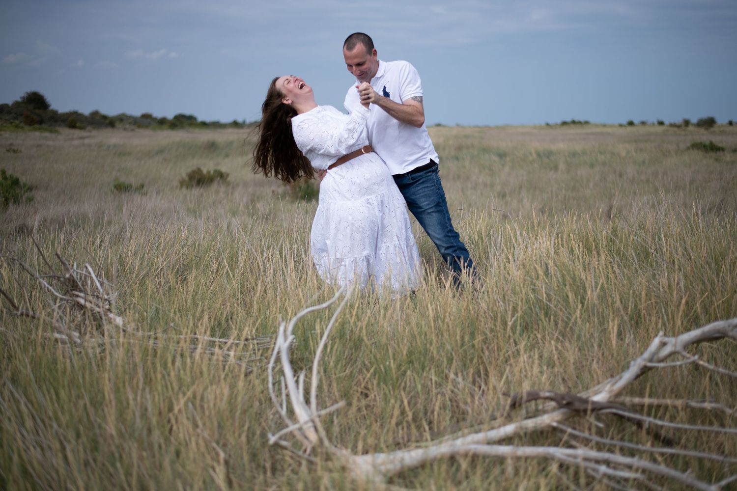 Couple dancing joyfully in a wide open grassy field, woman in flowing white dress with hair blowing in wind.