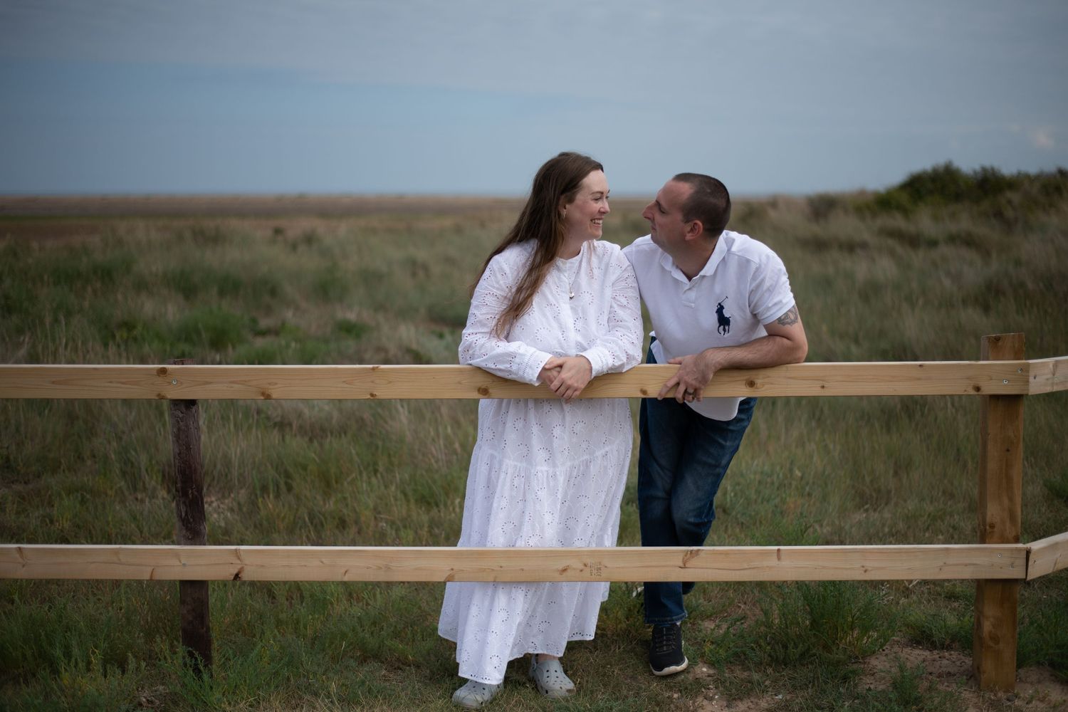 Couple leaning on wooden fence in open grassy field, sharing a romantic moment outdoors.