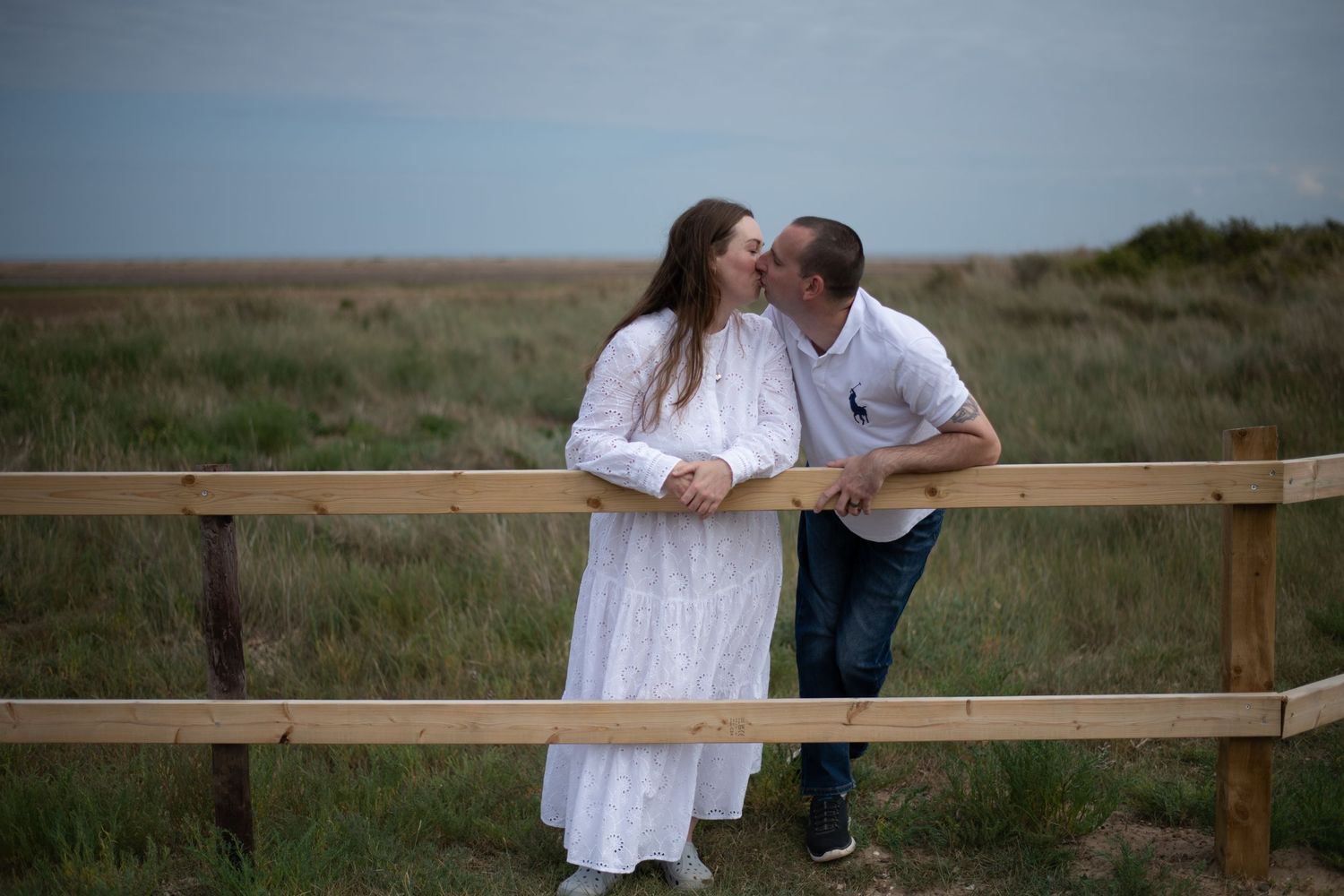 Couple sharing a romantic kiss by a wooden fence in an open grassy field, woman in white dress, man in white polo.