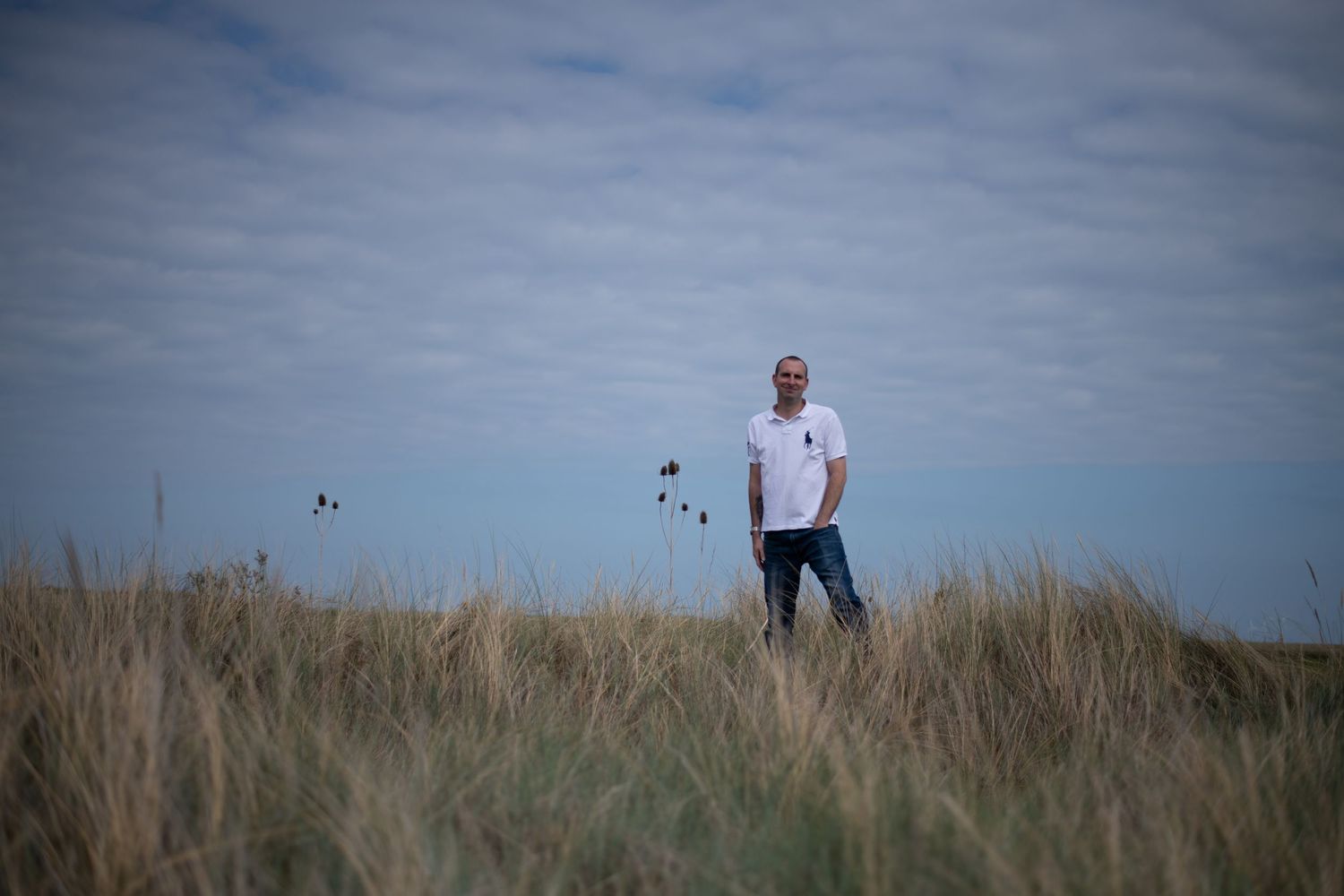 Man standing in coastal grass dunes under cloudy sky, wearing white polo shirt and jeans in scenic landscape.