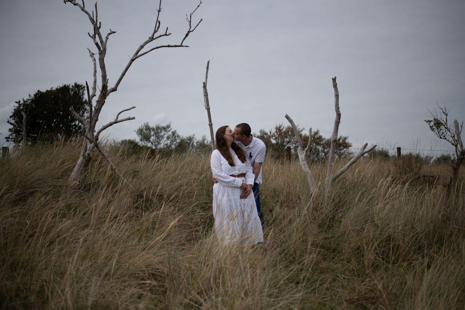 Couple sharing a romantic kiss in tall grass field with bare trees on a cloudy day, wearing white outfits.