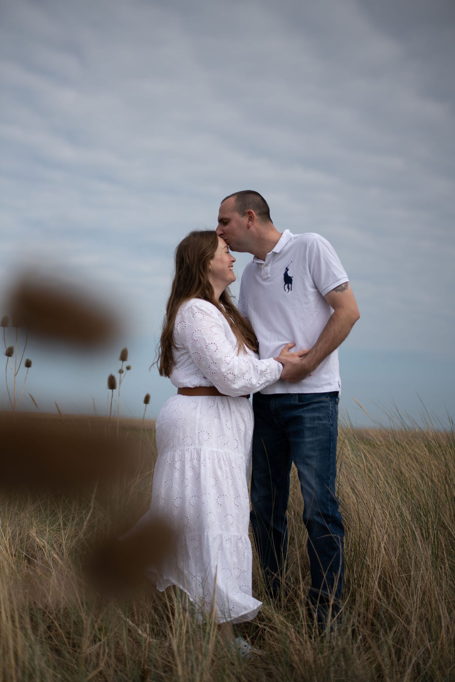 Couple sharing a romantic moment in golden grass field, woman in white dress and man in polo shirt embracing.