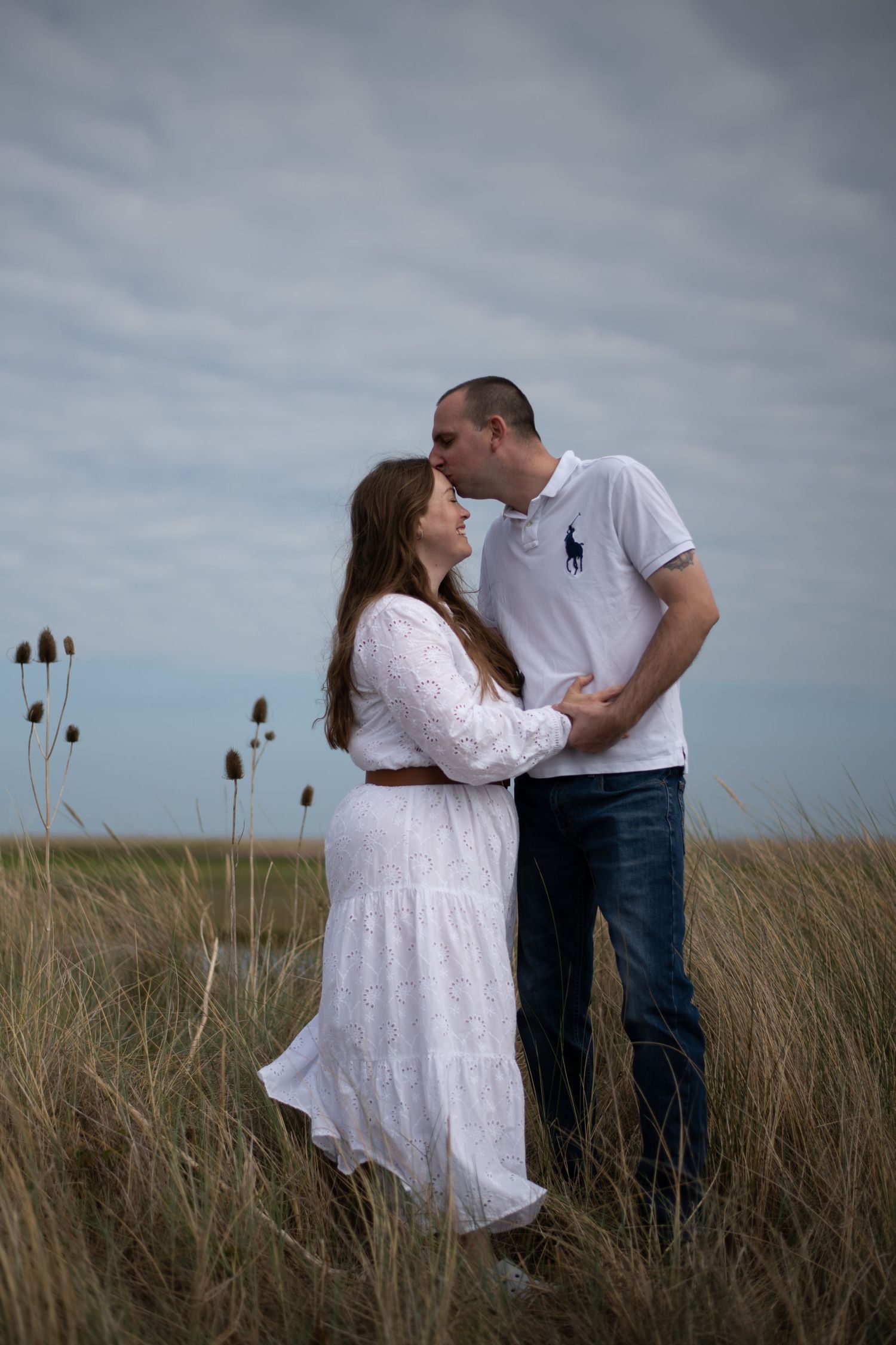 Couple sharing a romantic moment in tall grass field, woman in white dress, man in white polo shirt and jeans.
