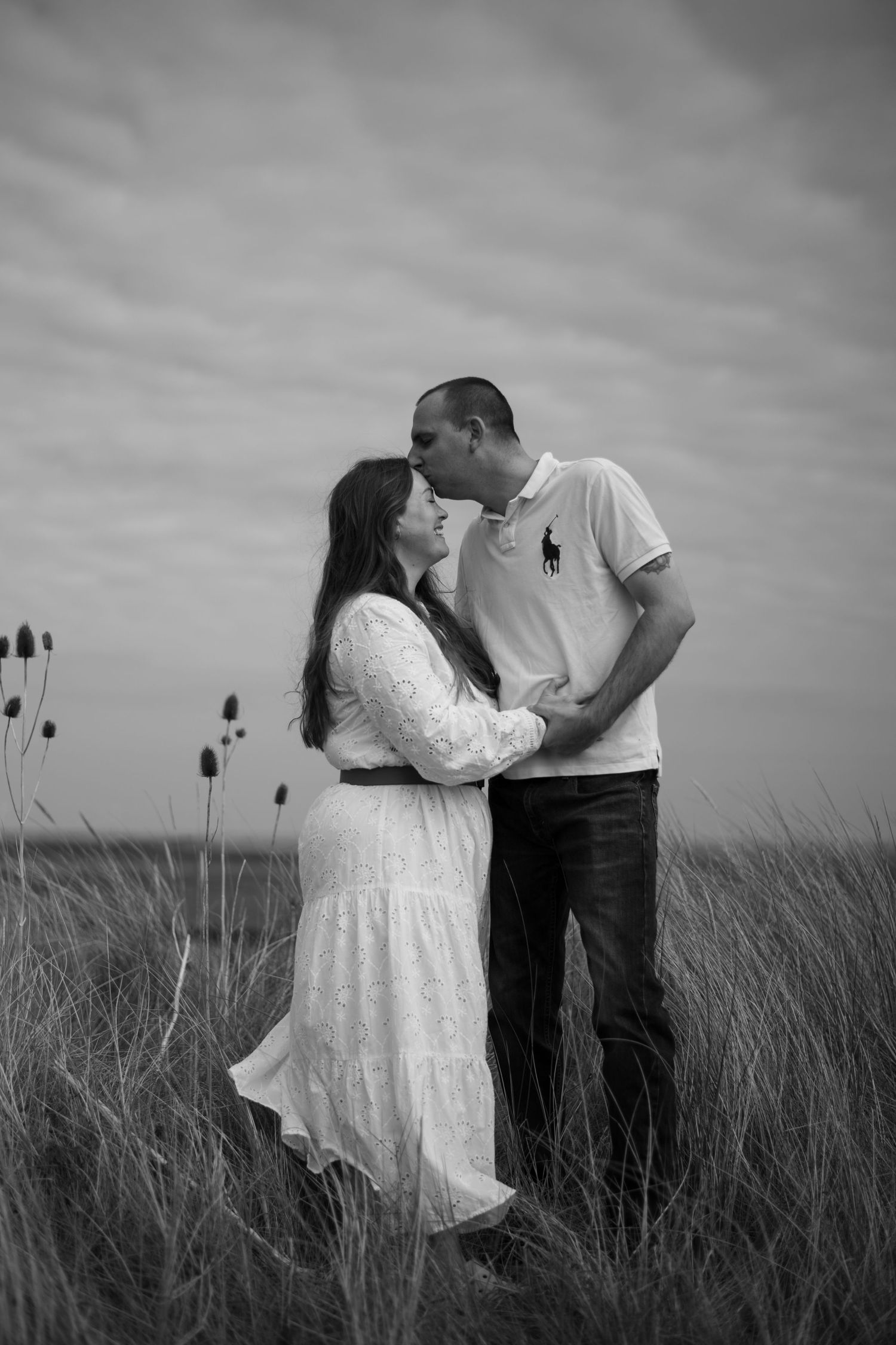 Black and white photo of a couple embracing and kissing in a grassy field under a cloudy sky.