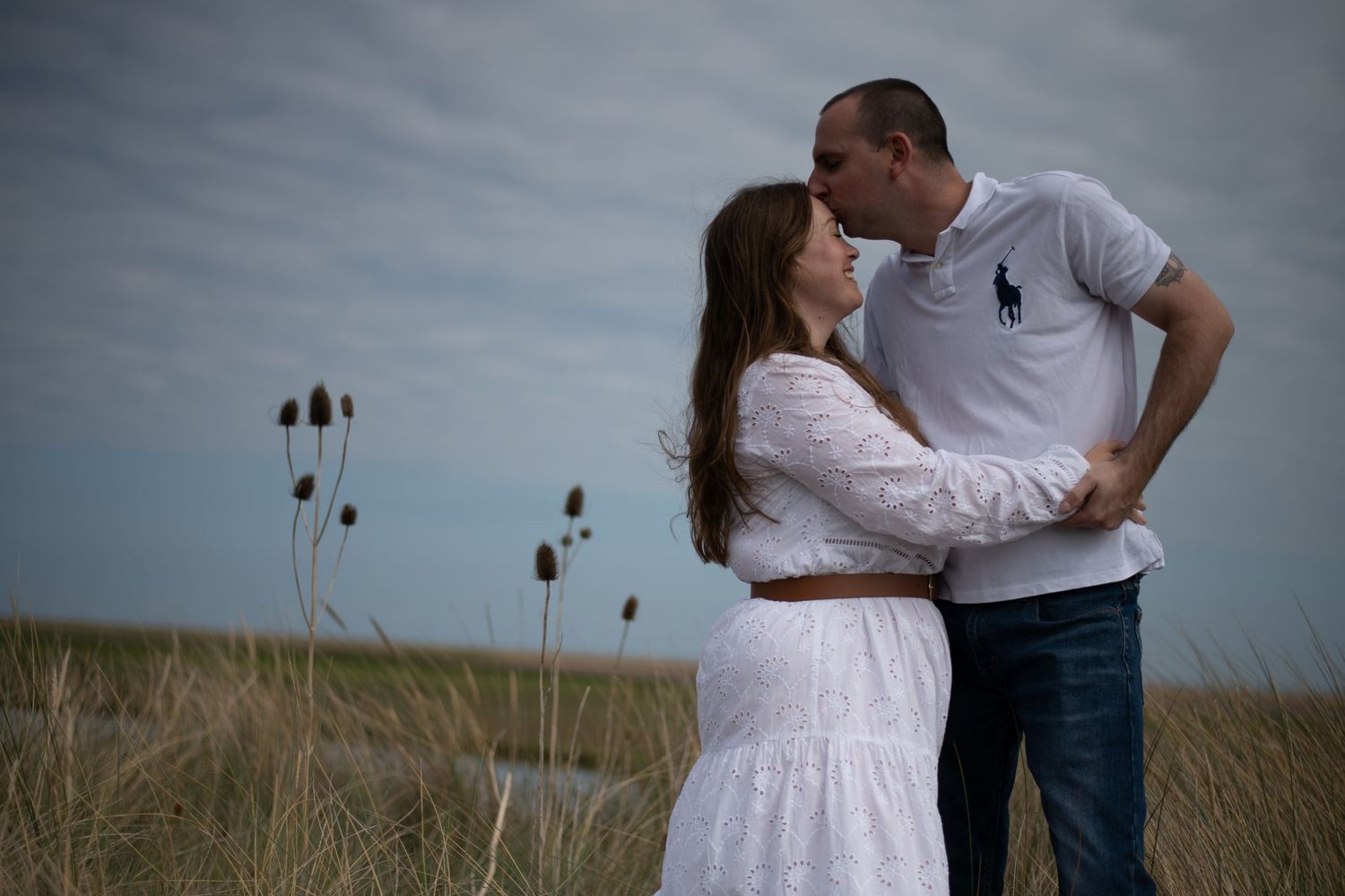 Couple sharing a romantic moment outdoors in a grassy field, woman in white dress, man in polo shirt, cloudy sky.