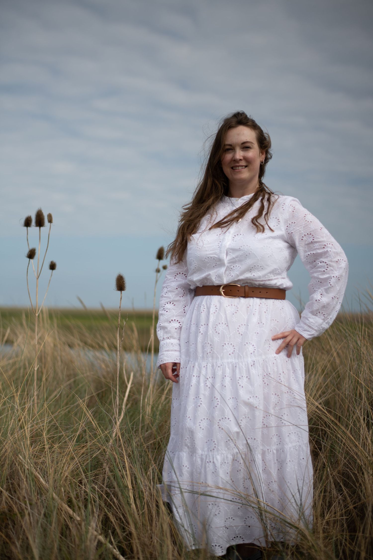 Woman in white lace dress with brown belt standing in tall grass field under cloudy sky.
