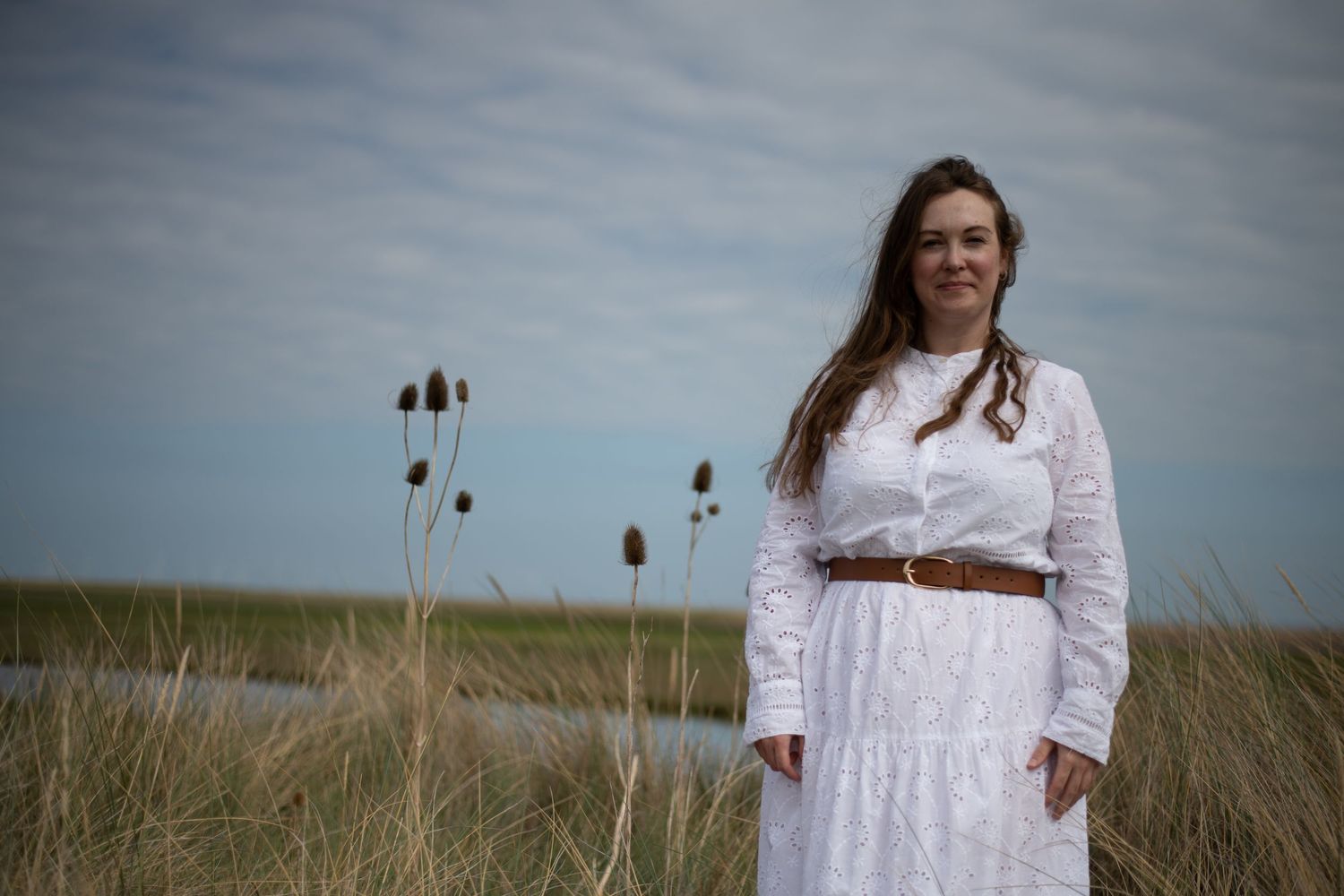 Woman in white lace dress with brown belt standing in coastal marshland.