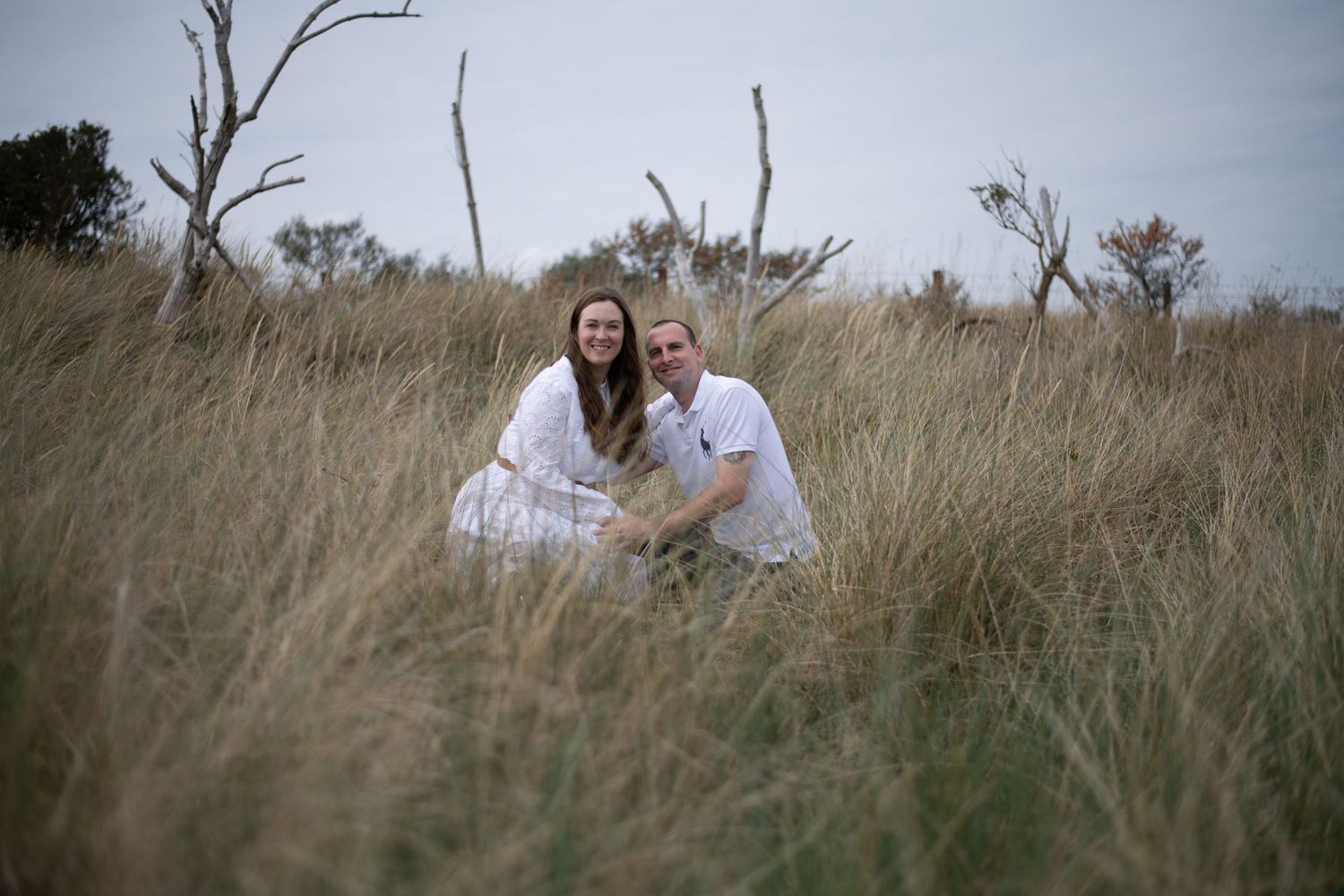 Couple in white outfits sitting together in tall grass field with bare trees in background.