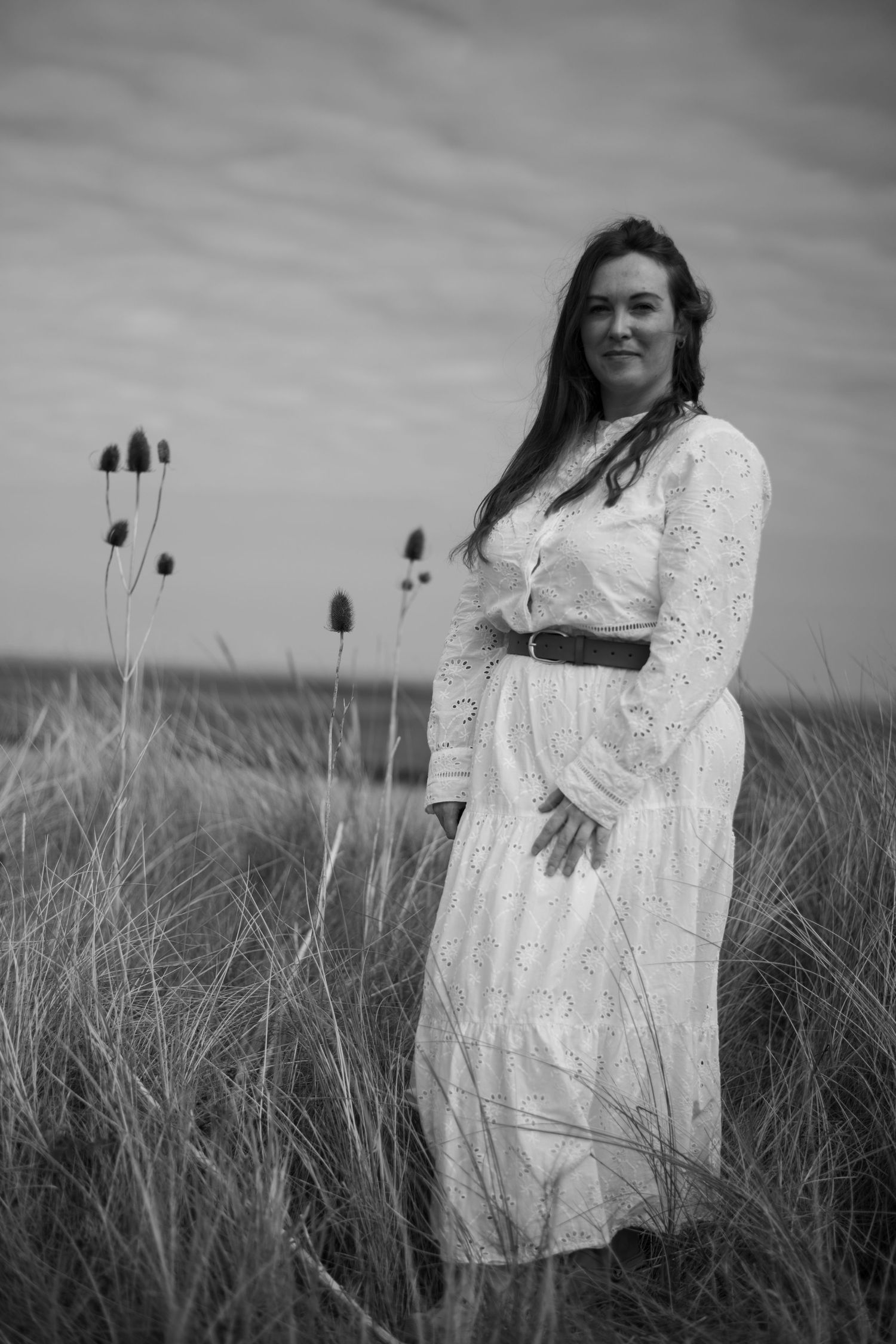 Black and white portrait of a woman in a white dress standing in a grassy field with wild teasel plants.