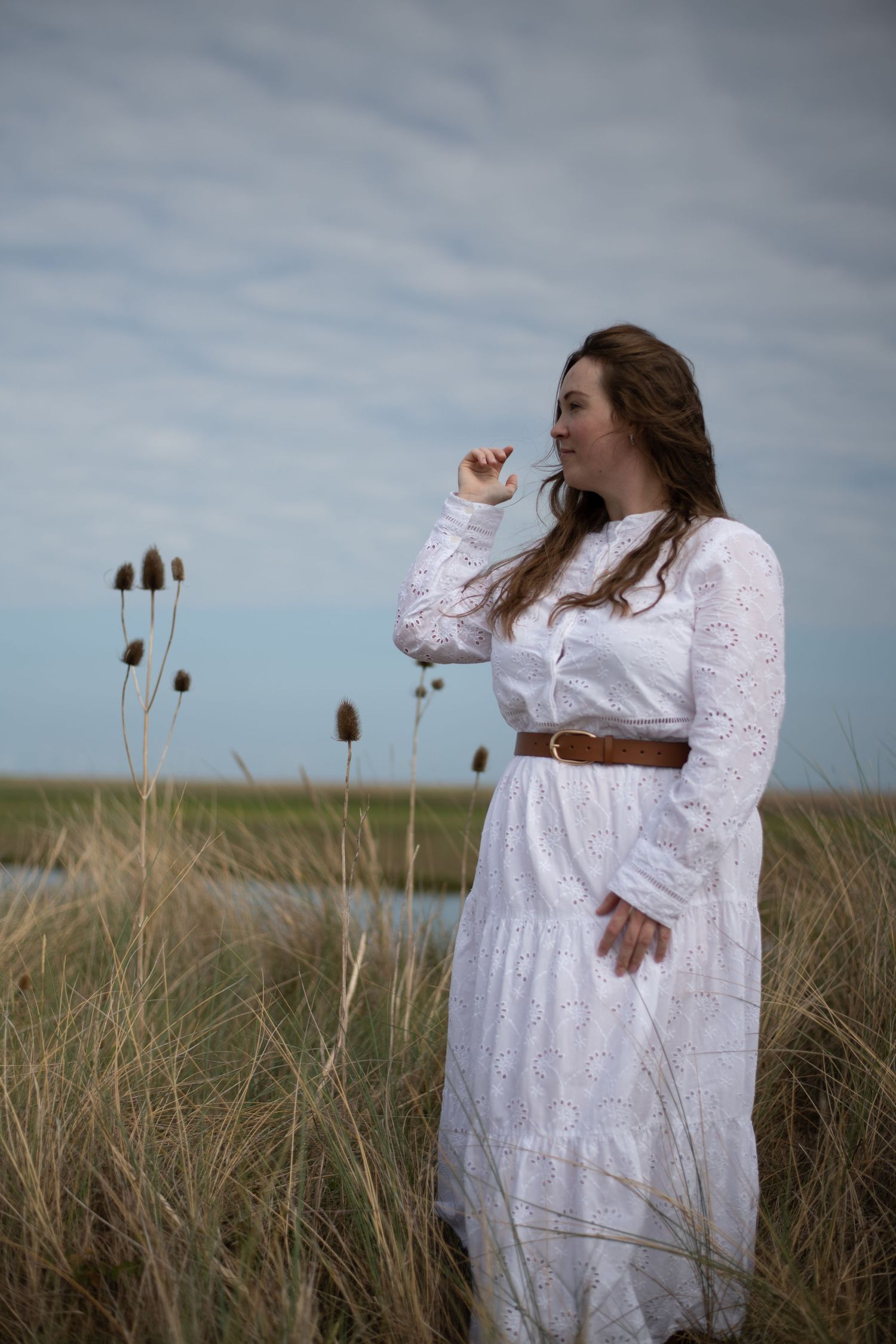 Woman in white lace dress with brown belt standing in marsh grassland under cloudy sky.