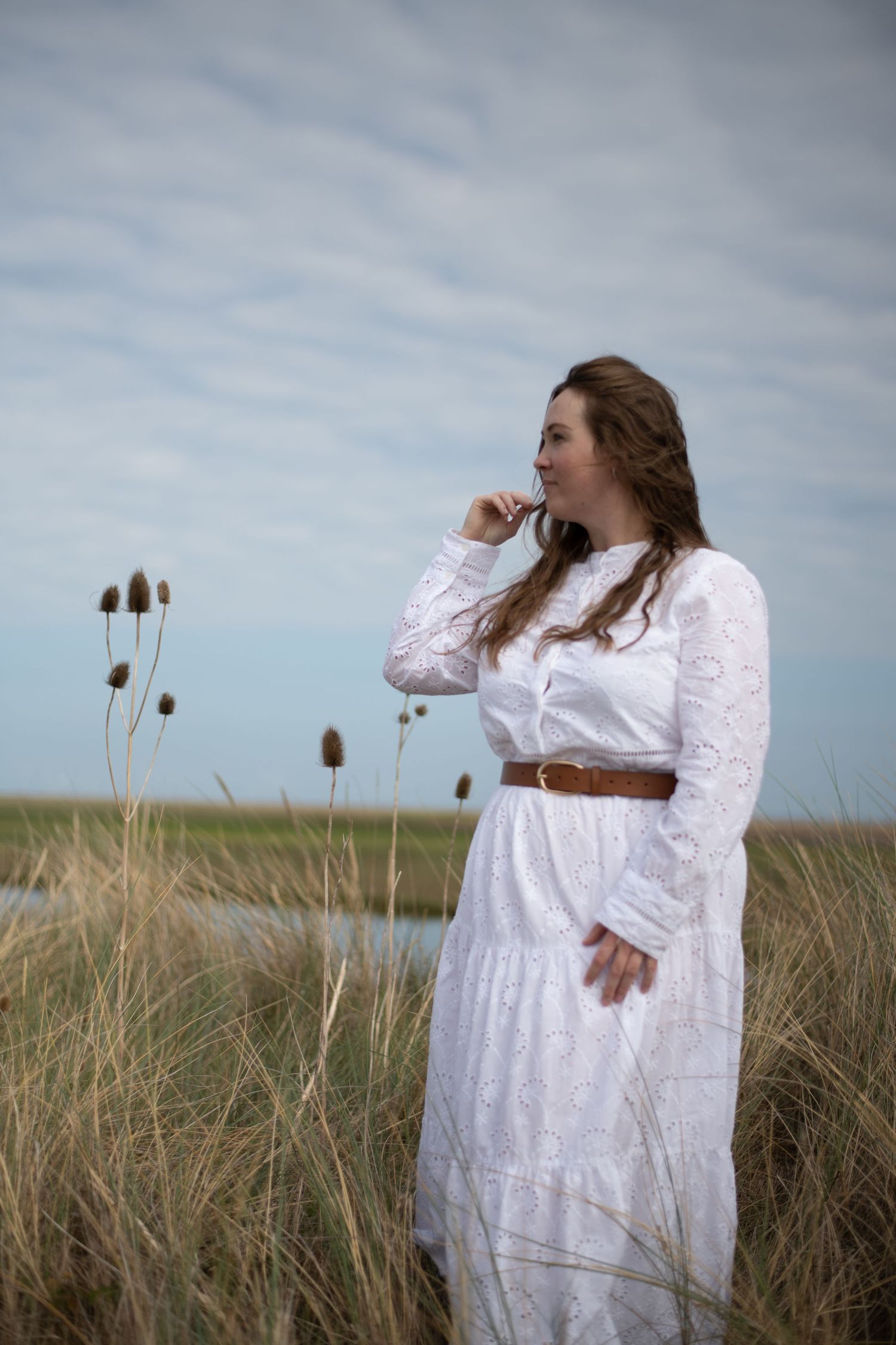Young woman in white lace dress with brown belt standing in marsh grass field under cloudy sky.