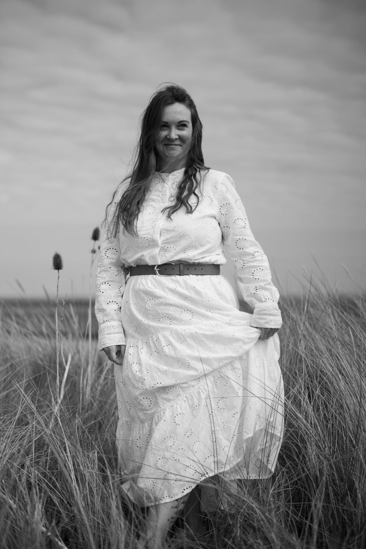 Black and white portrait of a smiling woman in a white lace belted dress standing in a grassy field.