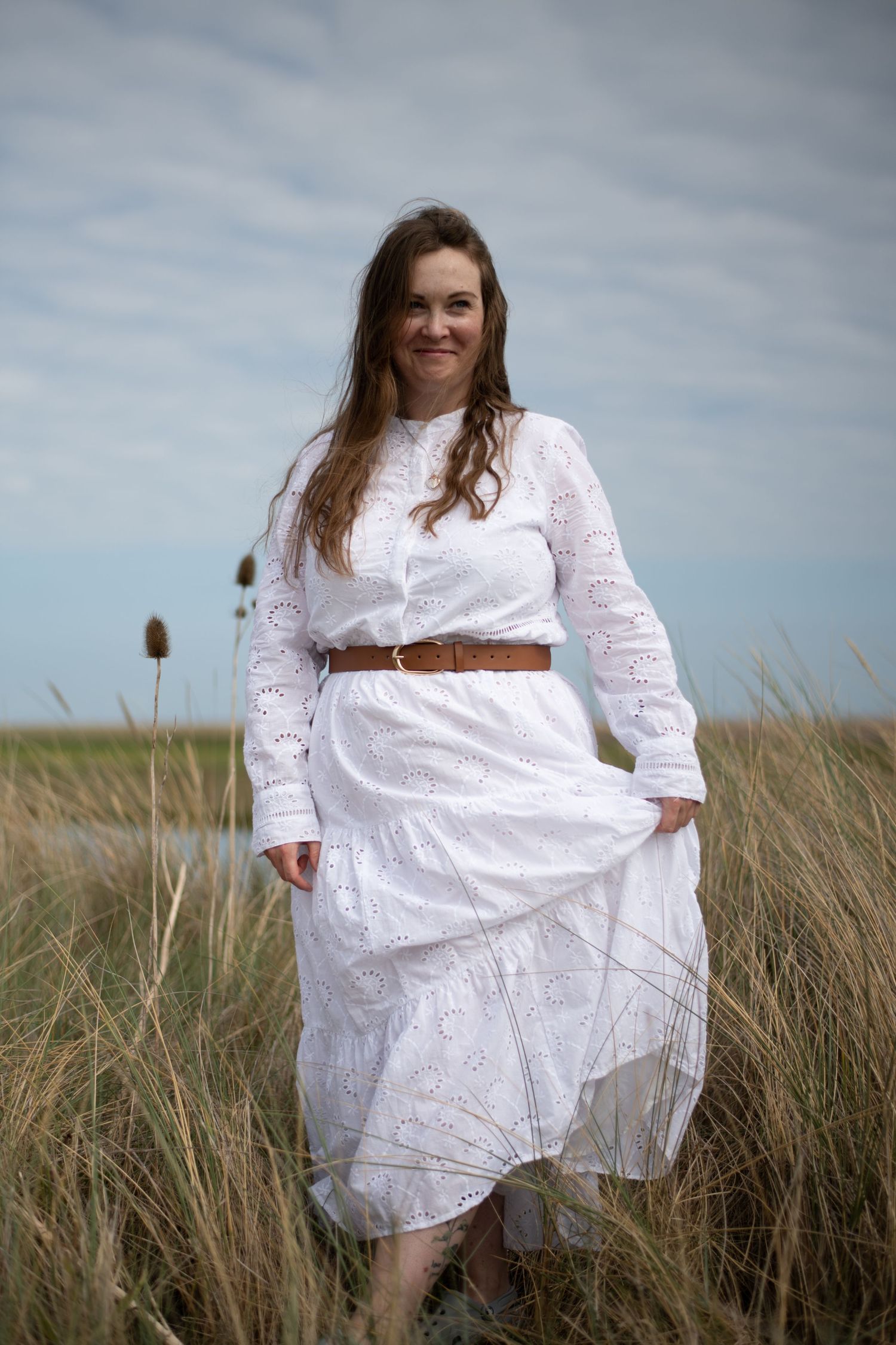 Woman in white lace dress with brown belt standing in a grassy field under a cloudy sky.