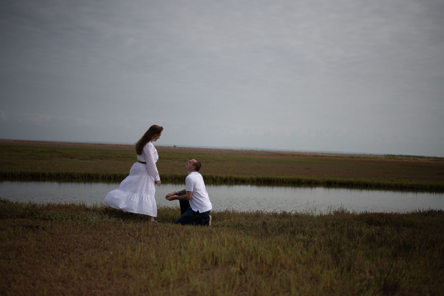 Couple in white attire near a serene marsh, woman standing while man kneels on grassy field.