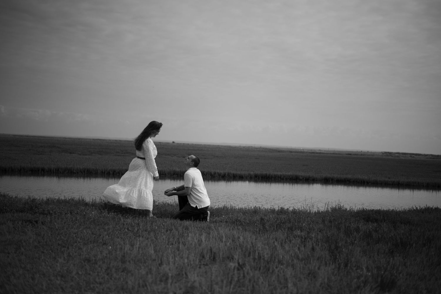 Black and white photo of a couple near marsh, man kneeling before woman in white dress.