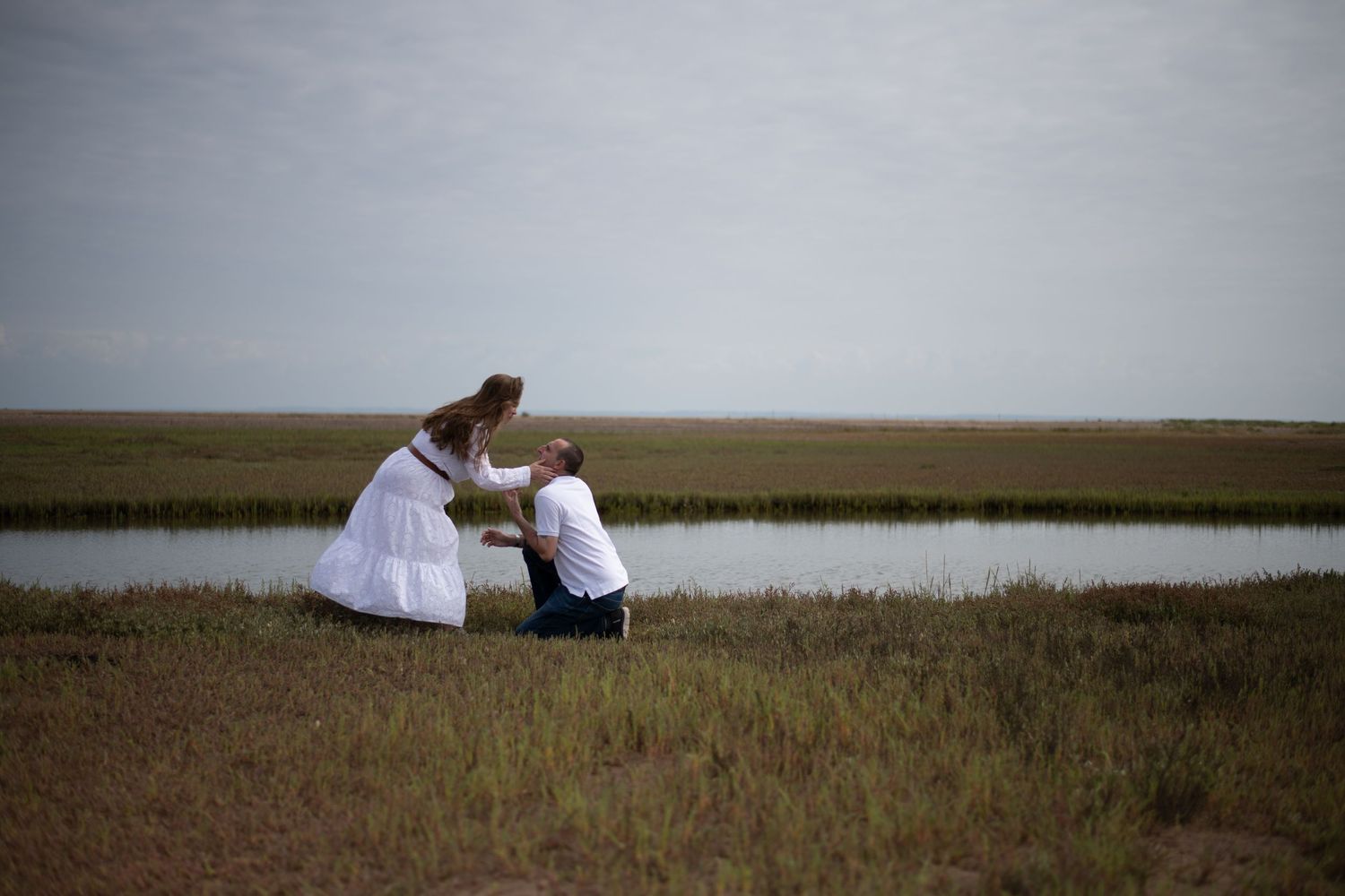Couple sharing a romantic moment by a marsh pond, man on one knee in a grassy field.