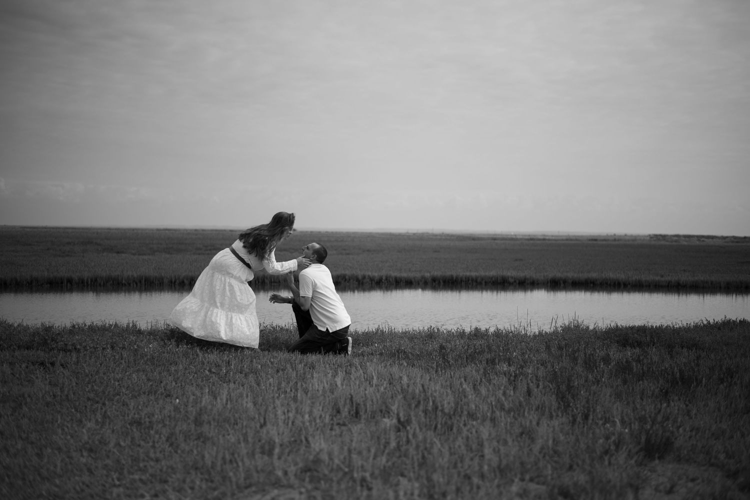 Black and white photo of a couple sharing a tender moment by a peaceful waterway in an open field.
