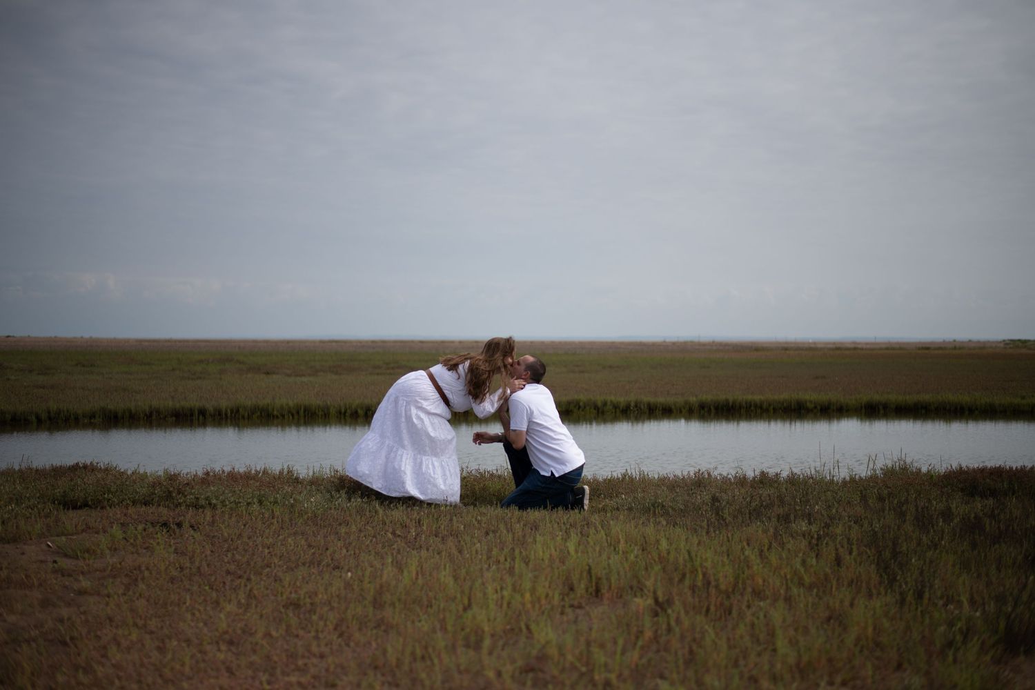 Couple sharing a tender kiss while kneeling in a coastal marsh field near a small pond.