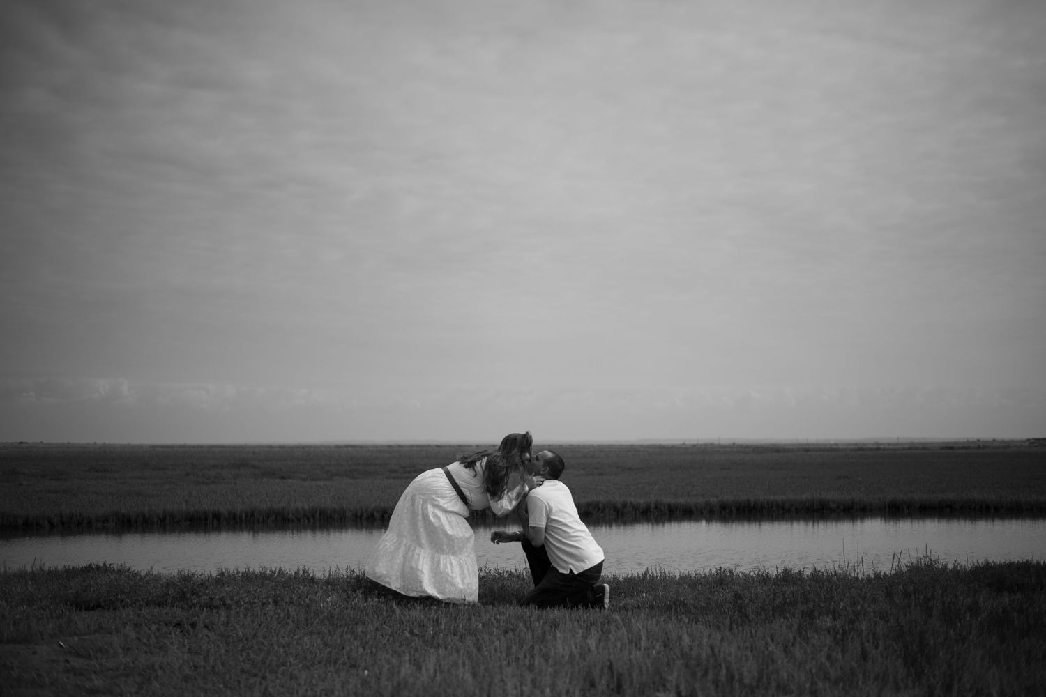 Black and white photo of couple sharing tender moment kneeling together by a peaceful marshland.