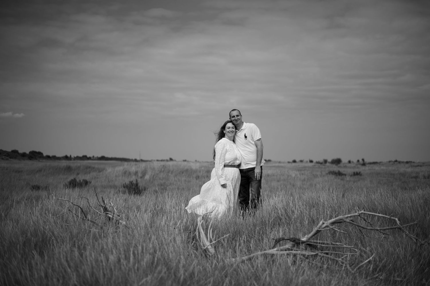 Black and white photo of a couple standing together in a vast open grassy field under a cloudy sky.