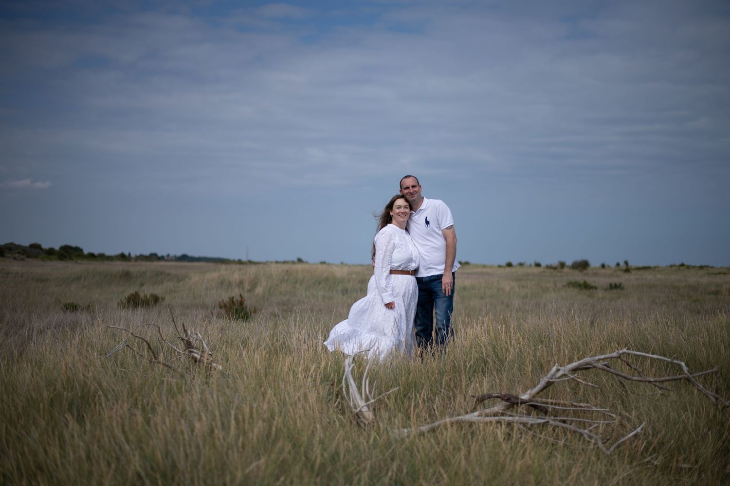 Couple posing in a grassy field, woman in white dress, man in jeans and white shirt, overcast sky.