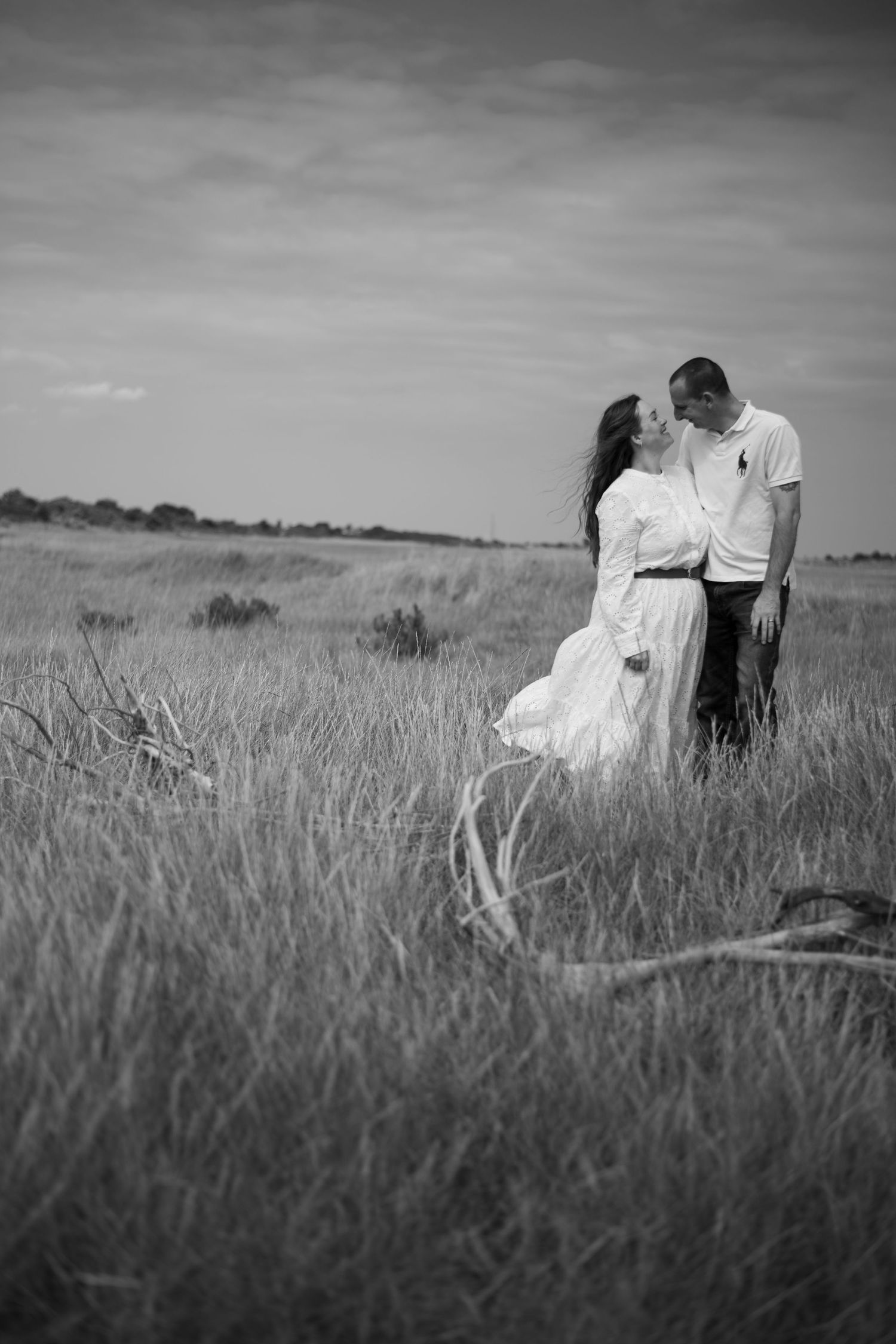 Black and white romantic photo of a couple kissing in a wide grassy field with driftwood nearby.
