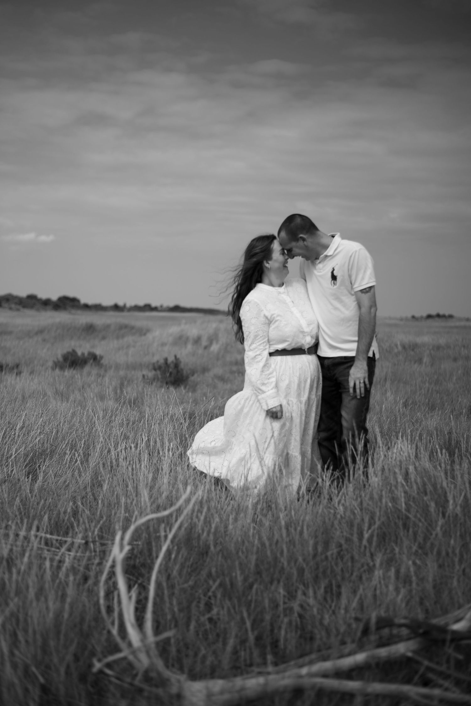 Black and white photo of a couple kissing in a vast grassy field under a dramatic sky.