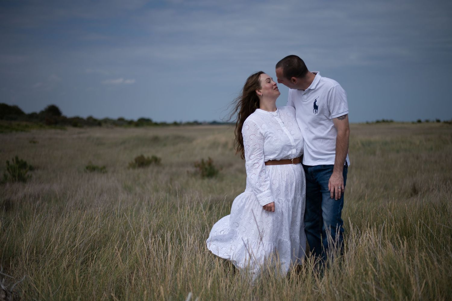 A couple shares a romantic kiss while standing in a vast grassy meadow under an overcast sky.