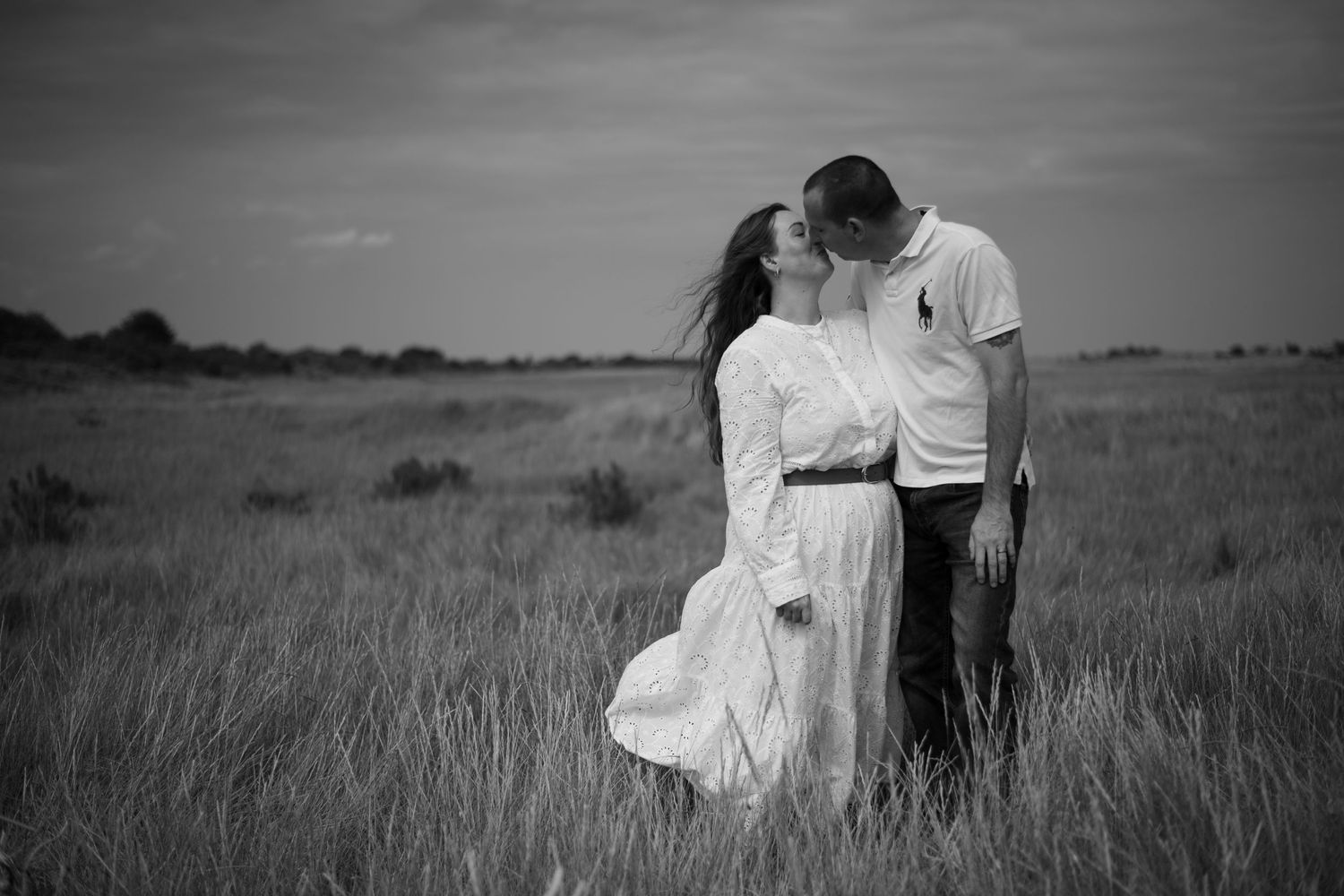 Black and white photo of couple kissing in a windswept grass field under dramatic sky.