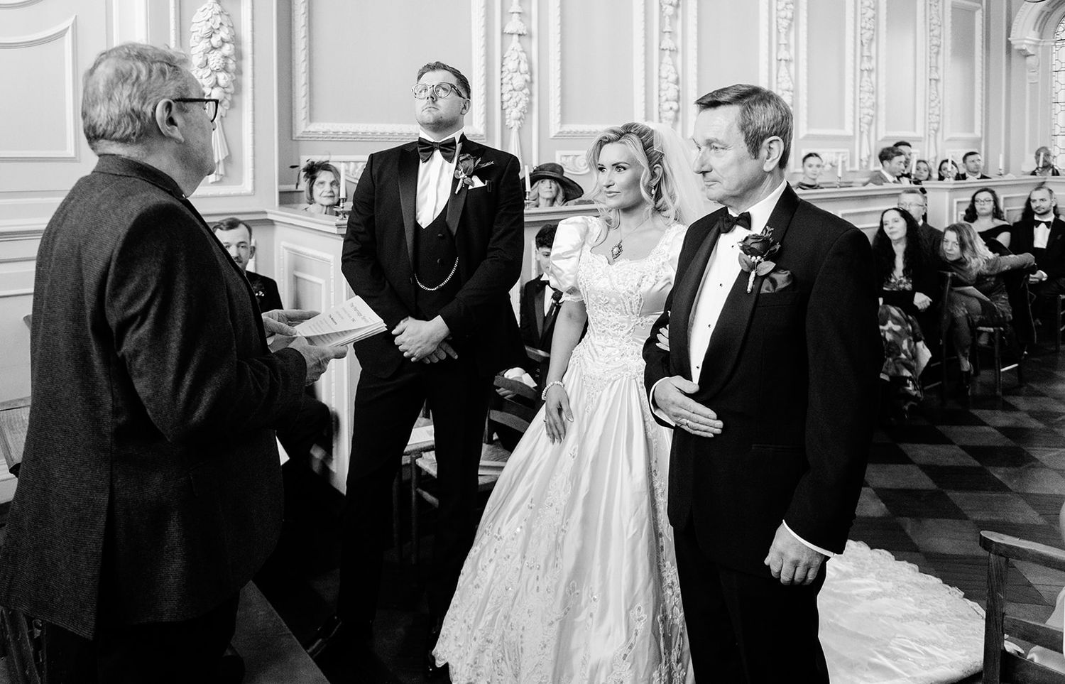 Black and white wedding ceremony photo with bride, groom, and officiant at elegant venue.