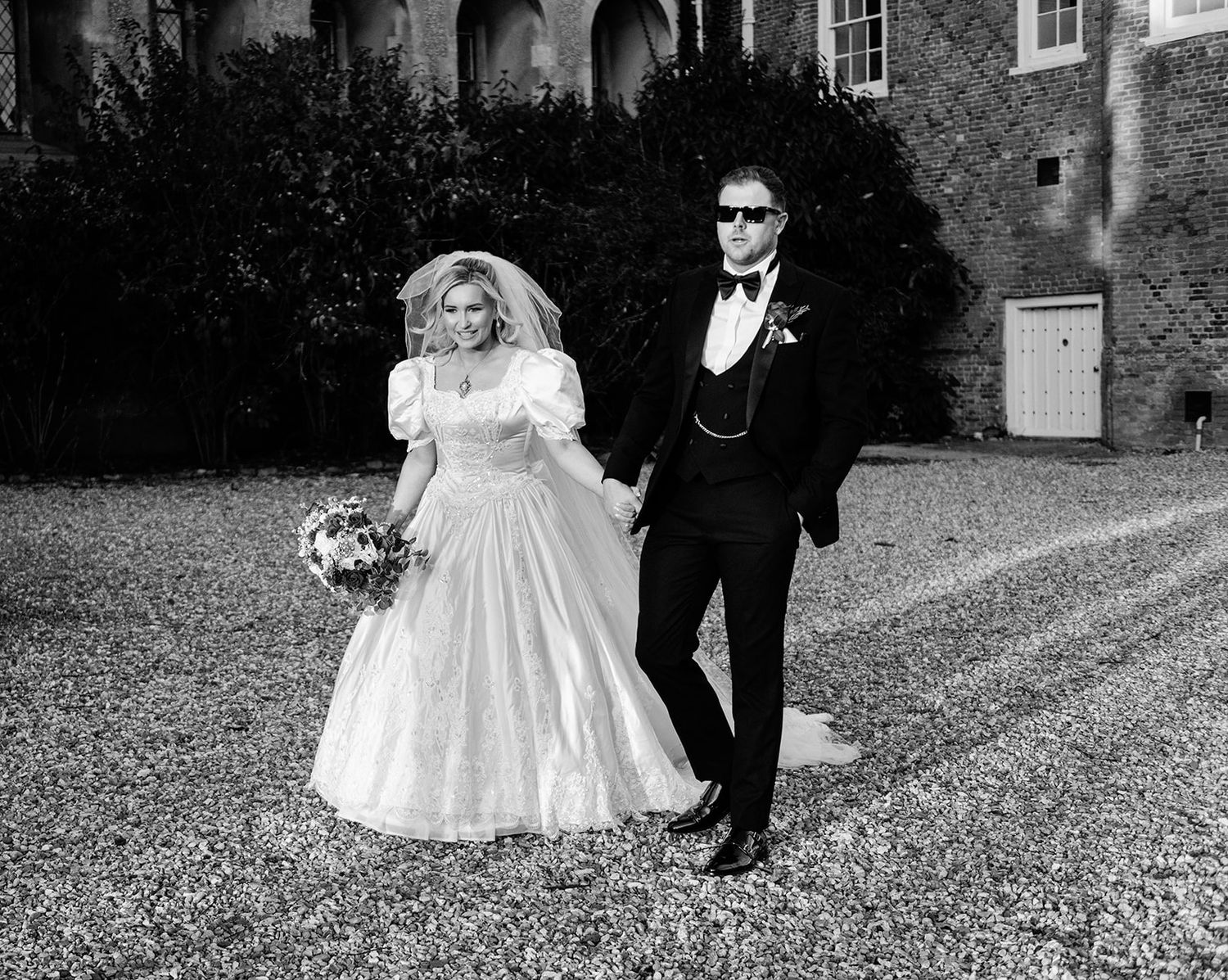 Black and white photo of a bride in a puffy wedding gown and groom in a tuxedo holding hands outdoors.