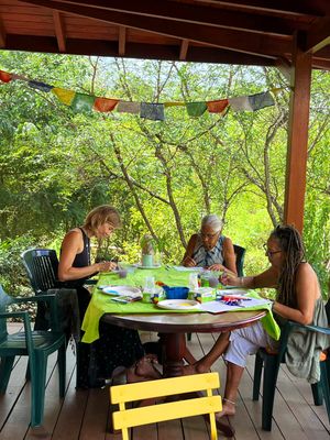 Three women crafting at a green table on a shaded outdoor porch surrounded by lush trees with colorful bunting flags.