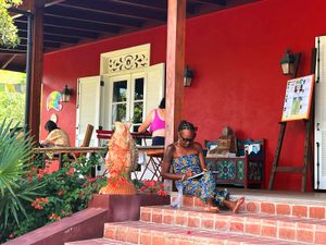 Woman reading on colorful red Caribbean porch with terracotta steps, tropical plants, and vibrant artwork displayed nearby.