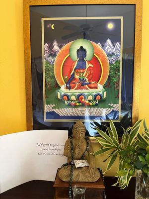 Tibetan thangka painting of Medicine Buddha in ornate frame above a stone Buddha statue with welcome card and green plant.