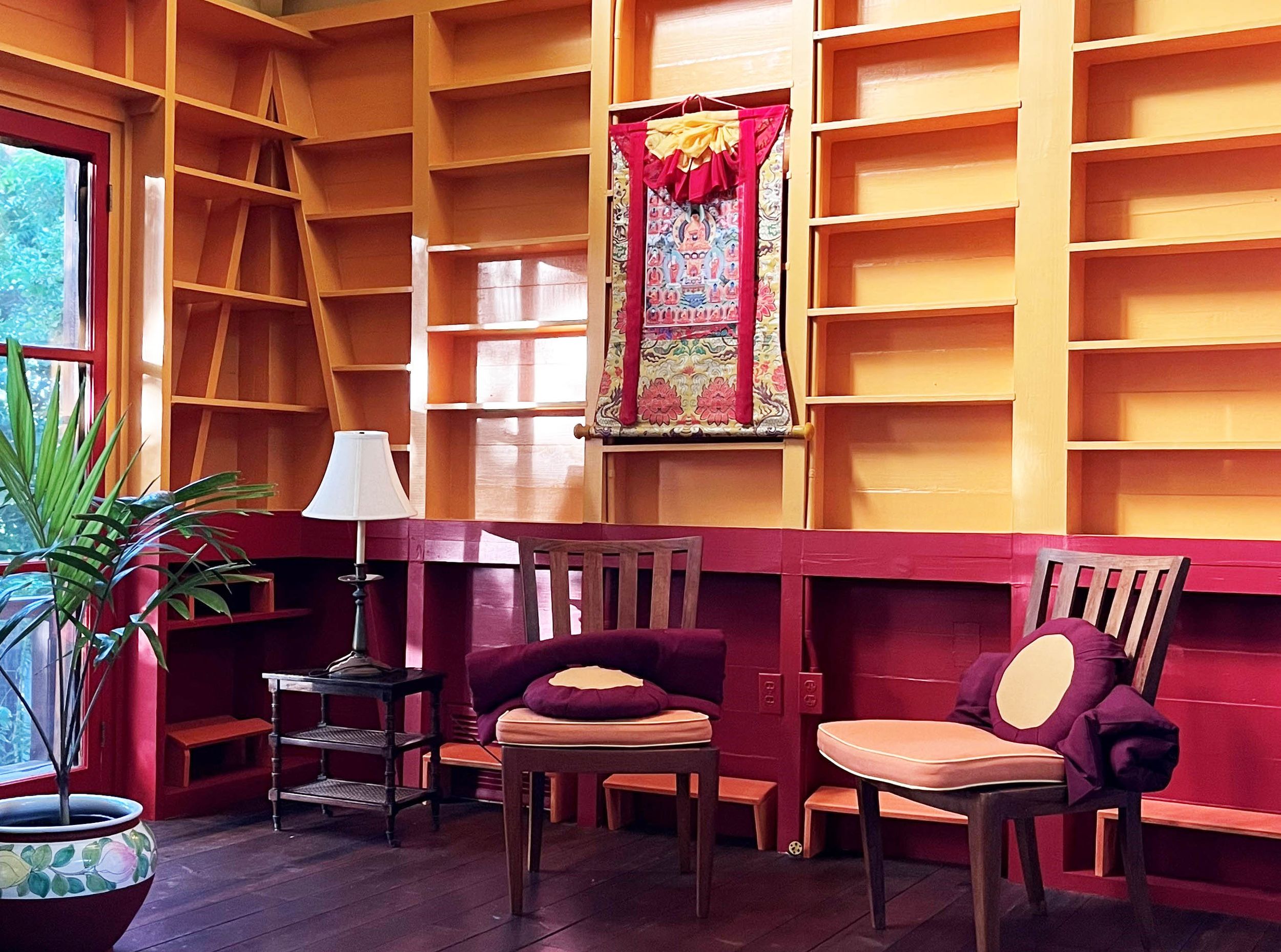 Cozy library room with yellow shelves, red walls, wooden furniture, a hanging tapestry, and a potted plant by the window.
