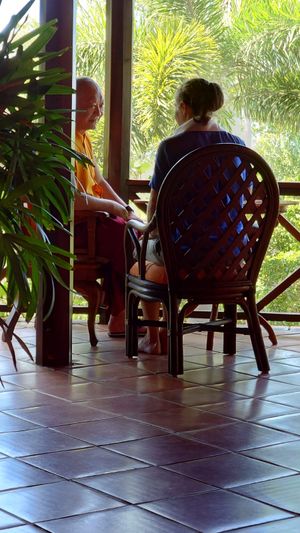 Two people sitting in wooden chairs on a tiled porch, conversing amid lush tropical greenery.