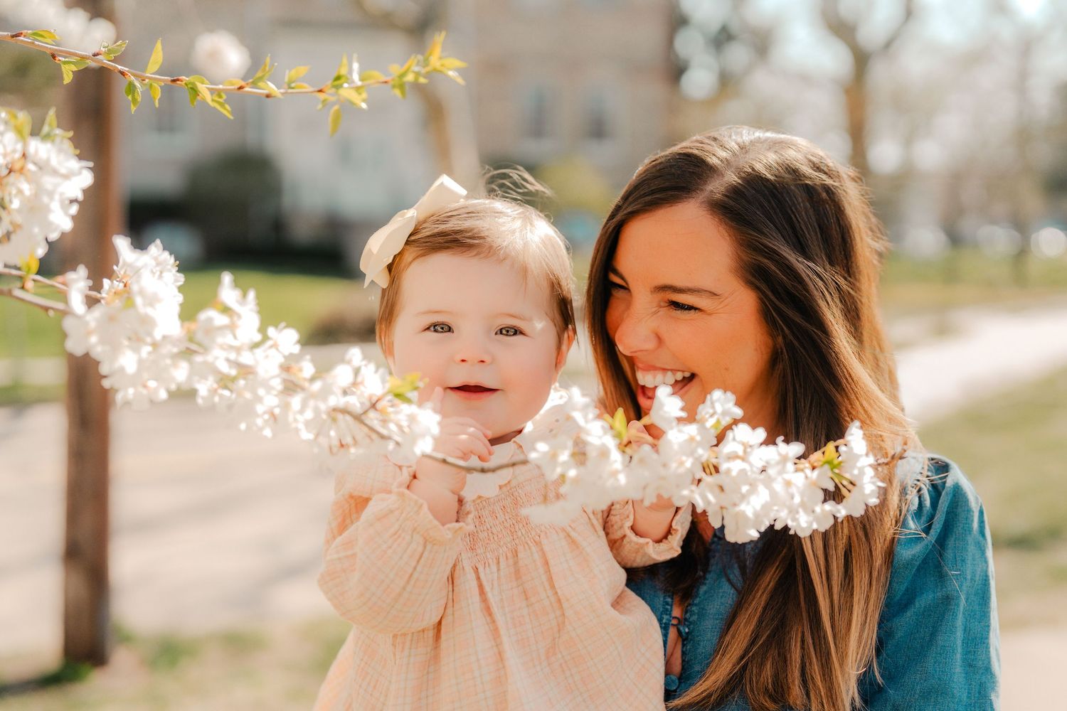 A smiling mother holds her happy baby girl near blooming white spring flowers outdoors.