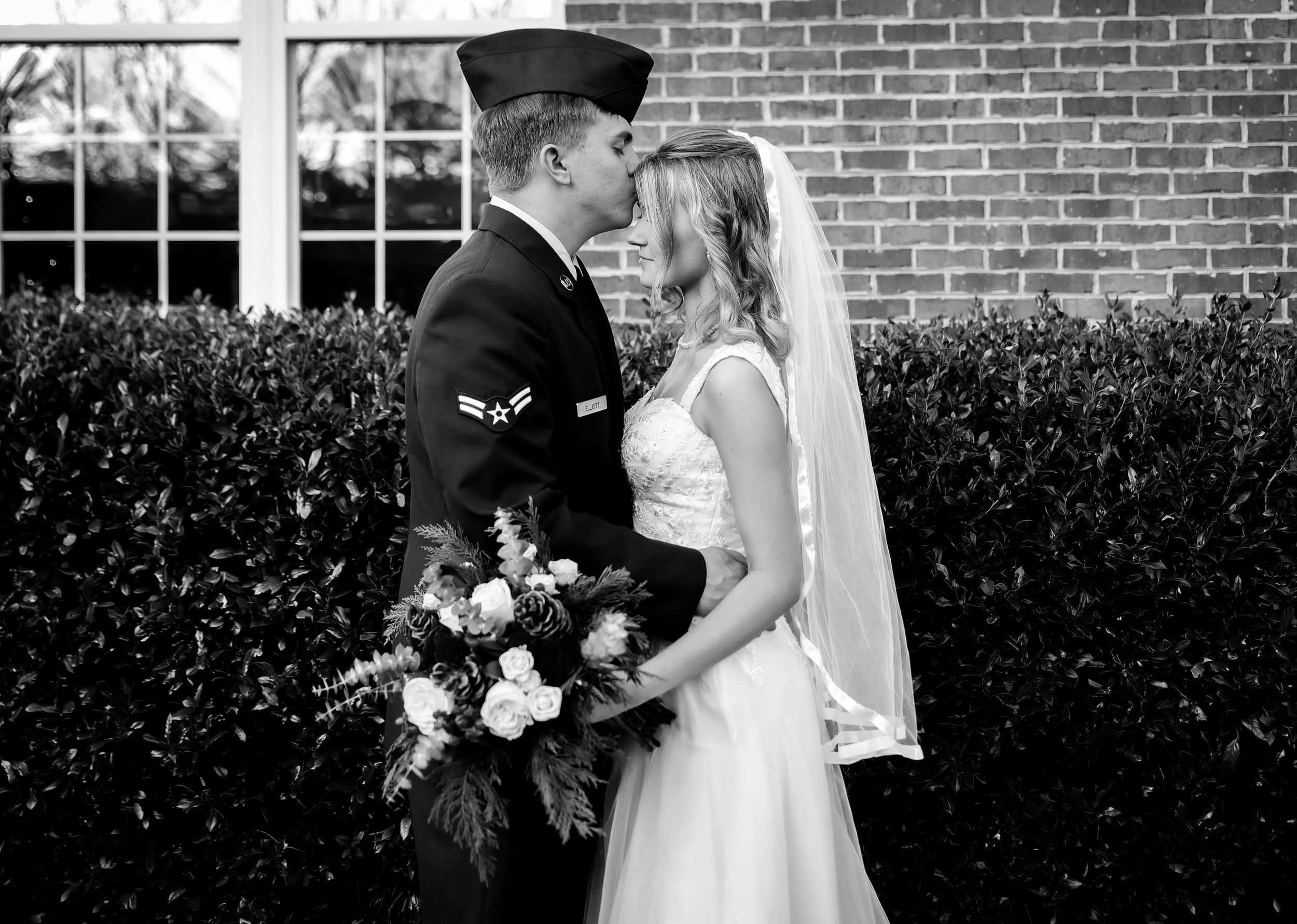 Black and white wedding photo of military groom in uniform kissing bride in white dress holding bouquet outdoors.