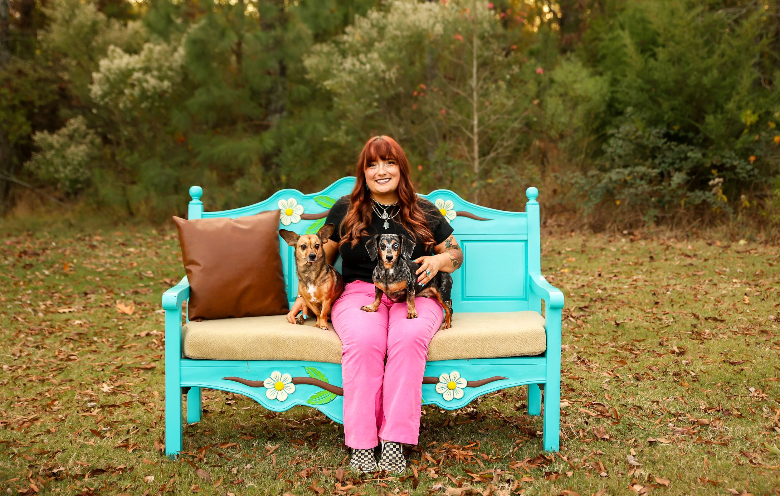 Woman in pink pants sits on turquoise bench outdoors with two small dogs amid autumn trees.