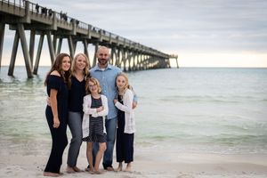 Family of five posing on a sandy beach near a pier with calm turquoise ocean waters in the background.
