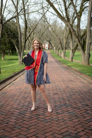 Smiling graduate in blue dress holds cap and wears red Chi Omega stole on brick path lined with trees.