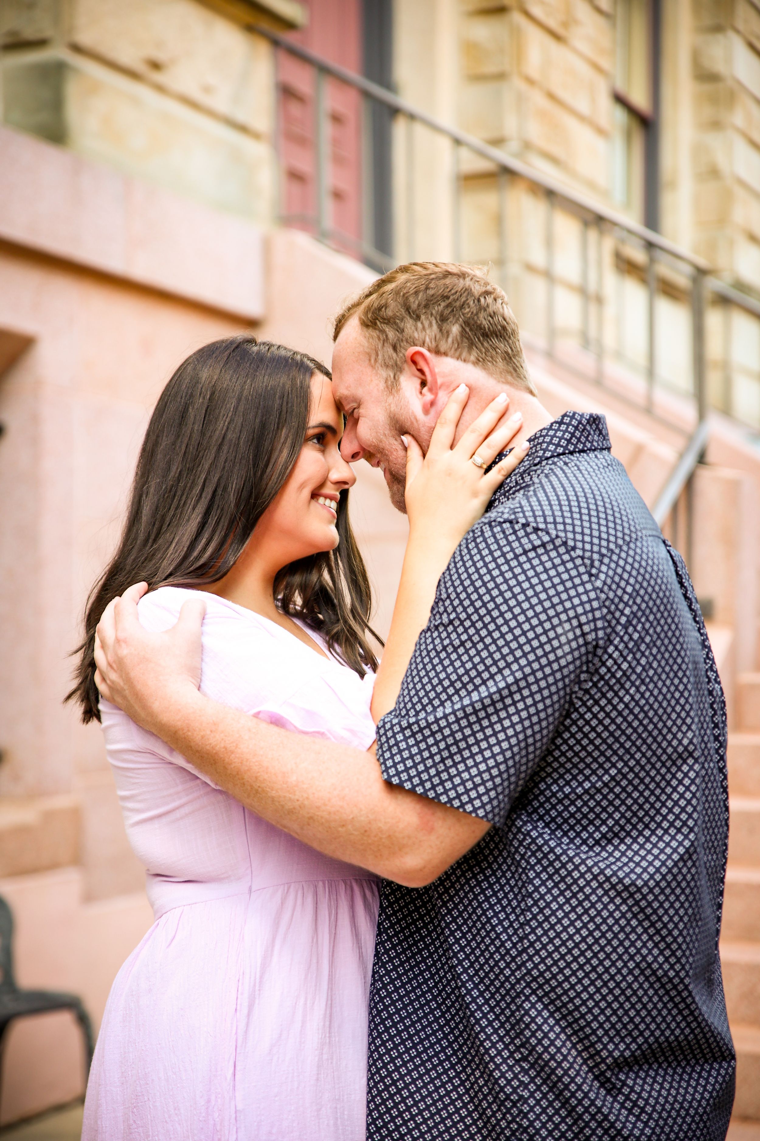 A couple shares a romantic forehead-to-forehead moment, the woman in a pink dress and man in a navy shirt, outdoors.
