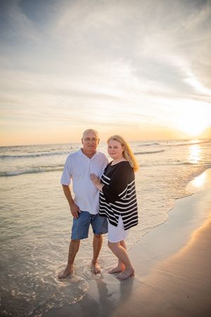 Couple standing barefoot on a sandy beach at sunset, waves gently washing ashore behind them.