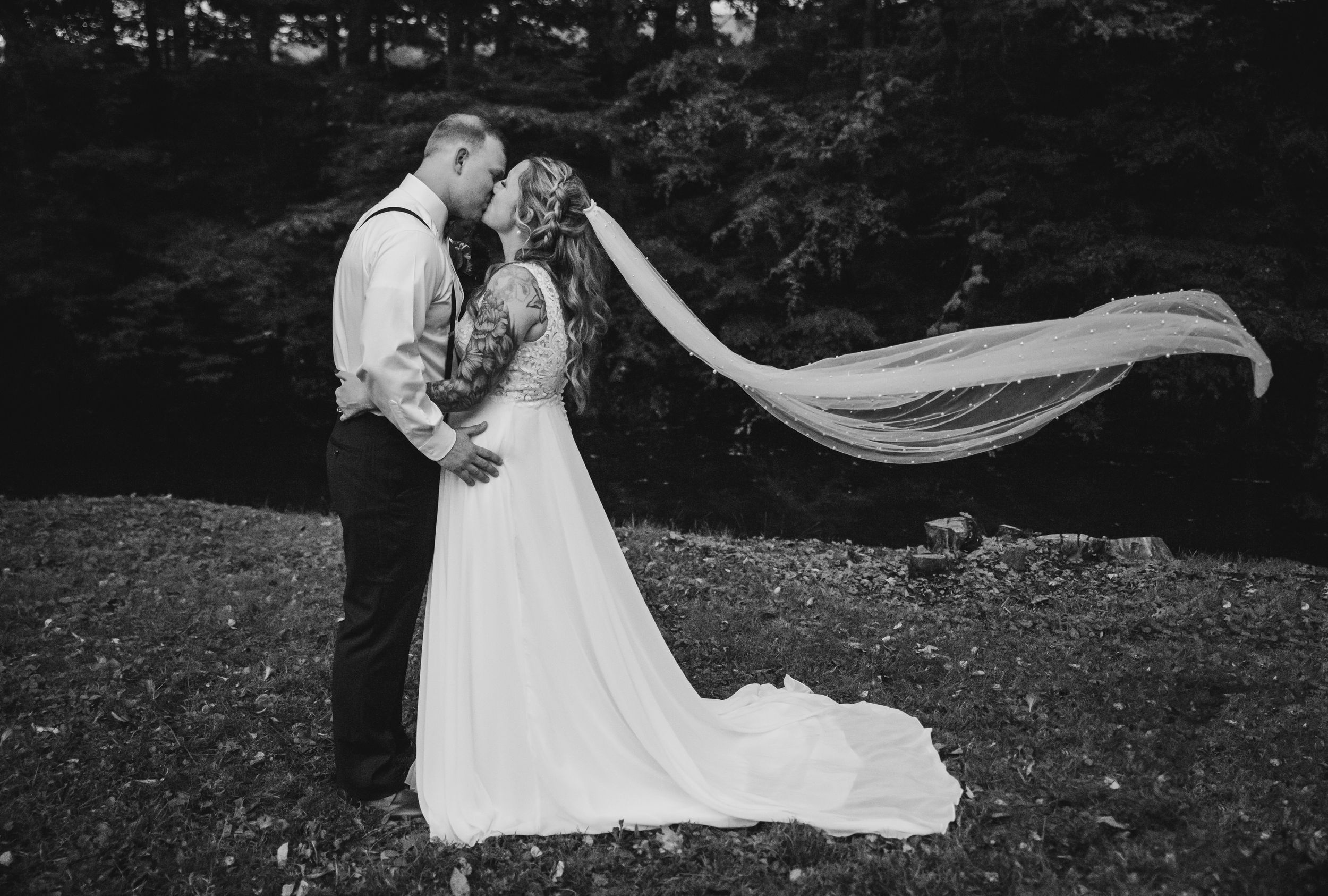 Black and white photo of a bride and groom kissing outdoors, with her veil flowing dramatically in the wind.