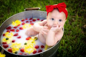 Baby girl with red bow sits in milk bath with strawberries and yellow flowers outdoors on green grass.