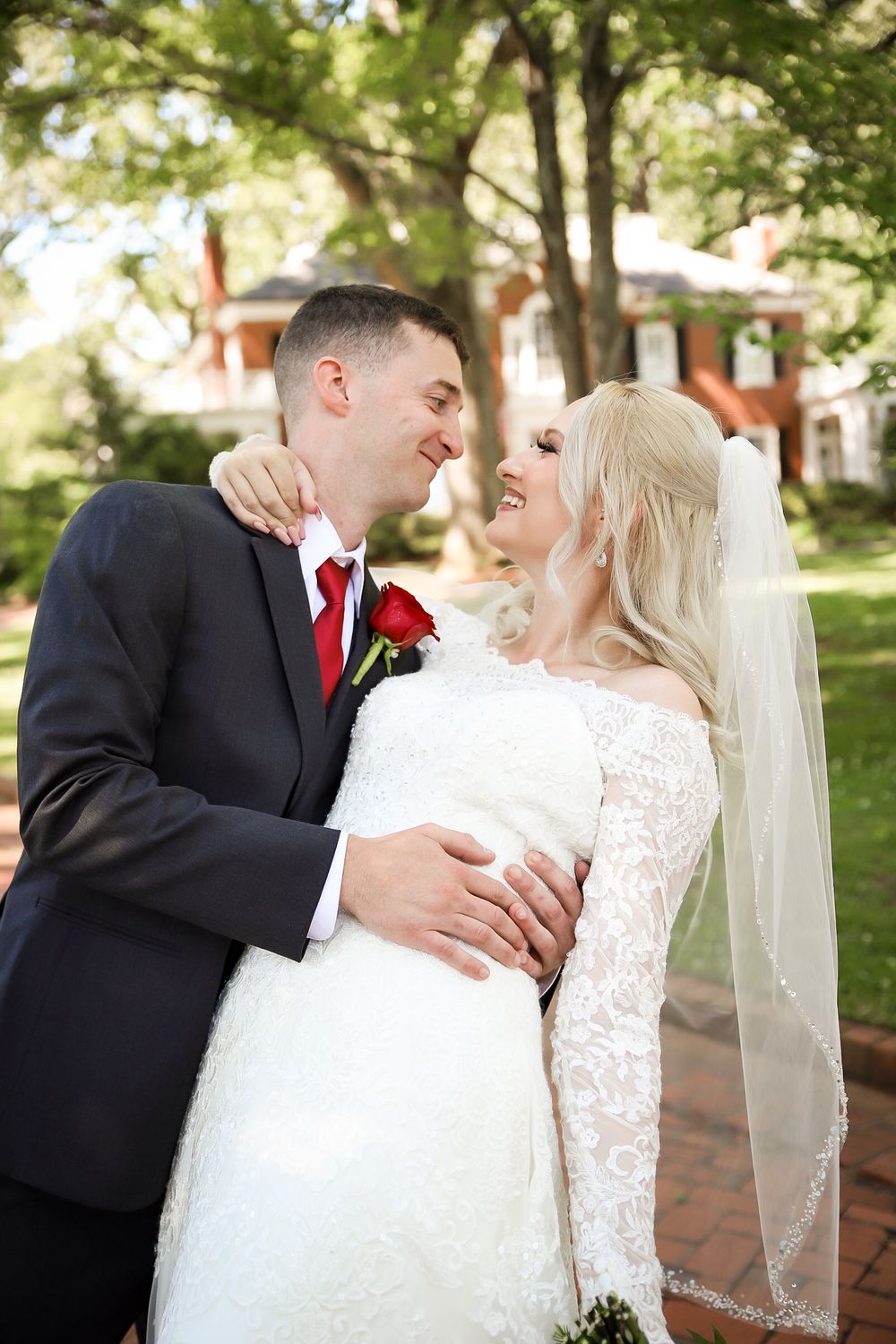 A bride and groom share a romantic moment outdoors, with the bride in a lace gown and veil, groom in a dark suit with red tie.
